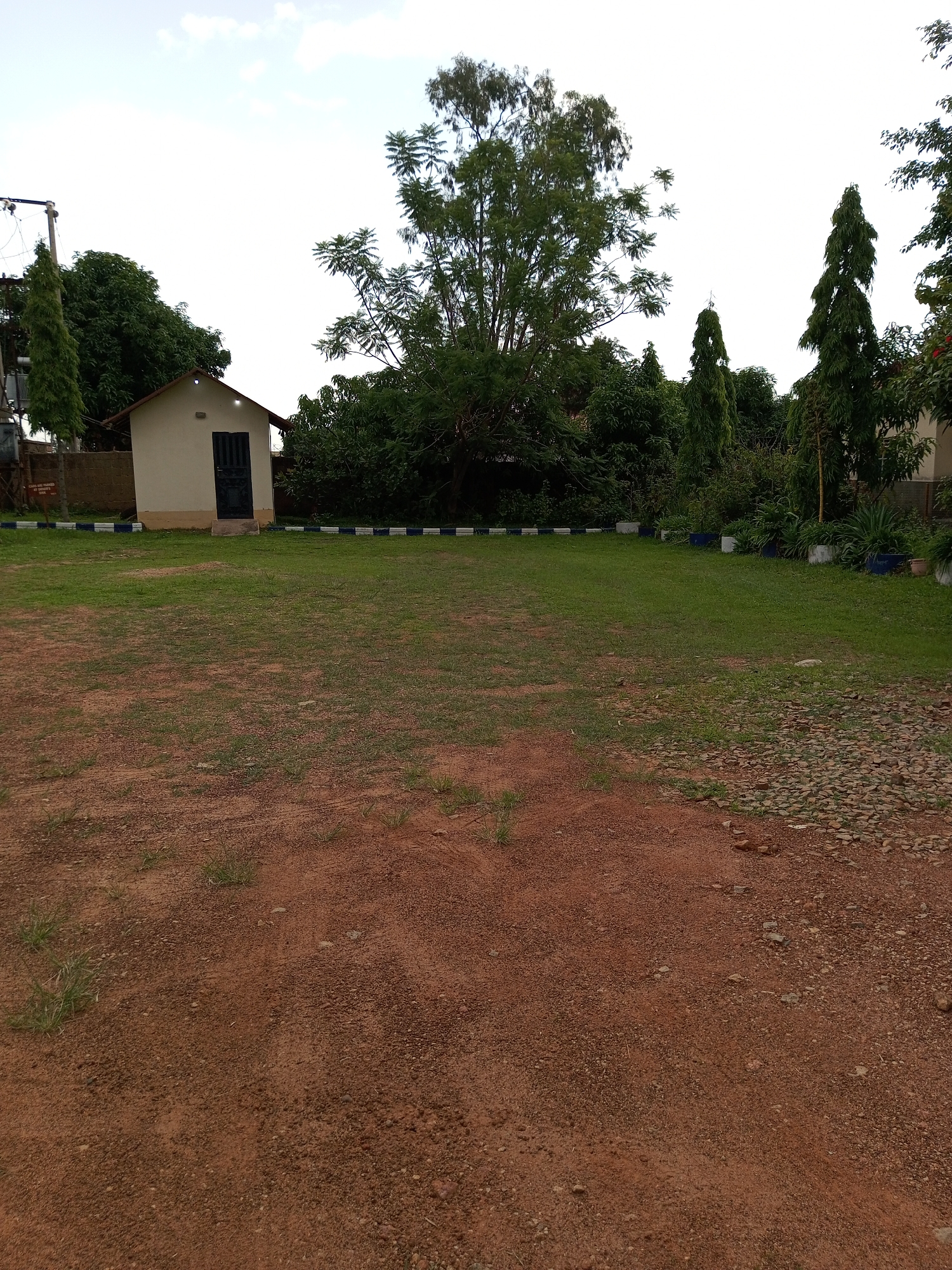 This is a photograph of a grassy area in front of a small building, likely a shed or outhouse. The building is light-colored with a dark door and sits behind a low decorative border of blue and white tiles. Several trees are visible in the background, including a tall, slender tree to the right and a fuller tree to the left. Some potted plants are also present. The ground in the foreground is a mix of red dirt and gravel with sparse patches of grass. The sky is overcast with a few clouds visible. The scene is likely located in Jos, Nigeria, as indicated by the surrounding vegetation and building style, which are common in the region.