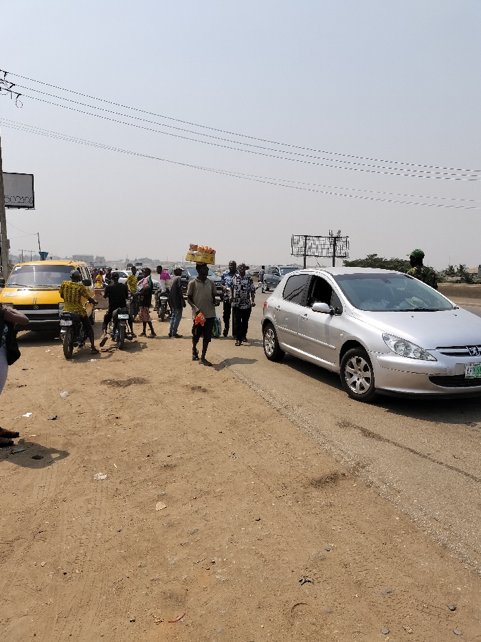The image depicts a roadside scene in Isheri, Nigeria, under clear, bright daylight conditions. The foreground features an unpaved, reddish-brown dirt shoulder marked with tire tracks and minor litter. To the right, a silver 5-door Peugeot 307 hatchback with a visible Lagos state license plate is positioned on a paved asphalt road, appearing stationary or moving slowly.
In the mid-ground, a dark-skinned male pedestrian, centrally located, walks from left to right. He wears a light-colored short-sleeved shirt and dark shorts, balancing a tray of bottled drinks or packaged snacks on his head while holding an item in his left hand. To his left, near an orange-yellow commercial bus (likely a Suzuki model), a group of several individuals, predominantly male and dressed in casual attire, are gathered on the unpaved ground. Two motorcycles, one with a rider, are also present in this area. Further into the background, additional vehicles are visible on the paved road, suggesting traffic.
On the far right, partially obscured by the silver Peugeot, a person in a dark green military or security uniform with a matching cap stands. Overhead power lines span the upper portion of the frame, and in the distant background, several large, empty metal billboard structures are visible against a hazy sky. A utility pole and a partial sign displaying "502100" are on the far left. The overall impression is that of a bustling urban or semi-urban roadside environment.