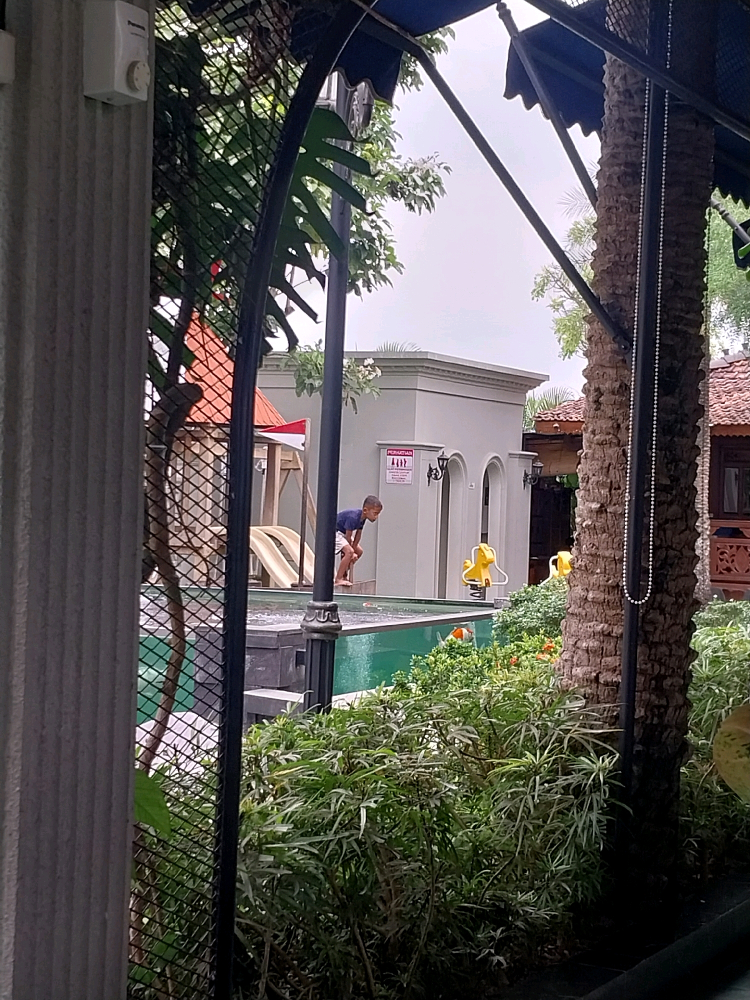 The image depicts a boy in a dark blue t-shirt and white shorts standing at the edge of a swimming pool in Bugisan, Indonesia. He is leaning forward, appearing to be looking into the water. The pool has a dark gray border and is filled with clear green water. To the left of the pool, a playground structure with two yellow slides is visible, situated behind a dark metal fence. A building with arched doorways and a red-tiled roof is in the background, with a sign displaying a red and white flag. The environment is lush with green foliage surrounding the pool and playground. A tall palm tree with a textured trunk stands to the right of the frame, partially obscuring a beaded string hanging down. The sky is overcast.