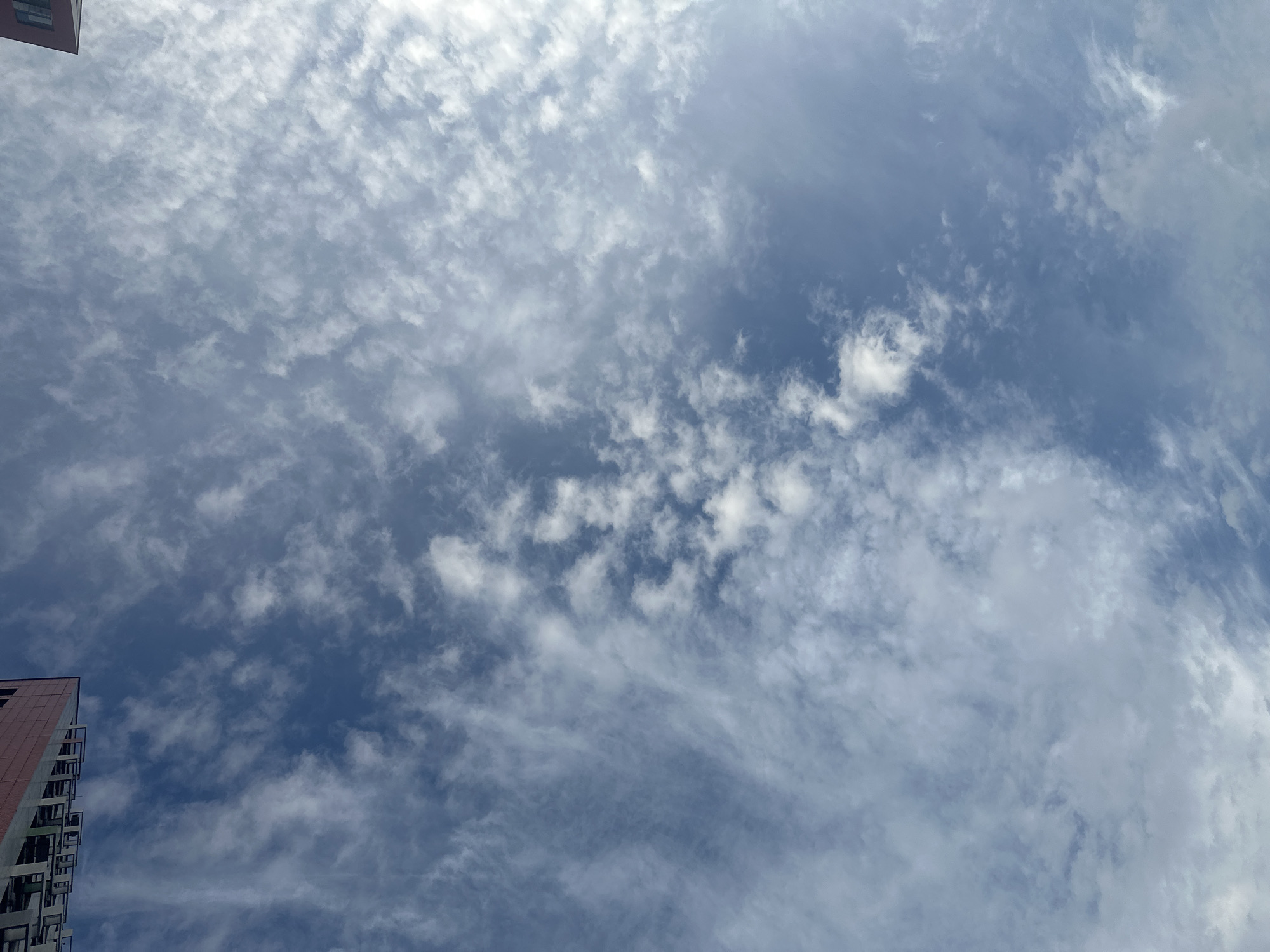 The image is a low-angle shot of a cloudy blue sky, viewed from ground level. In the upper left corner, a portion of a multi-story building with balconies and red siding is visible. In the upper right corner, another building with a dark roof and window is partially visible. The clouds are scattered and wispy, with some appearing more dense than others. The scene is likely an urban environment, given the presence of buildings, but no specific location within Moscow, Russia can be determined from the image.