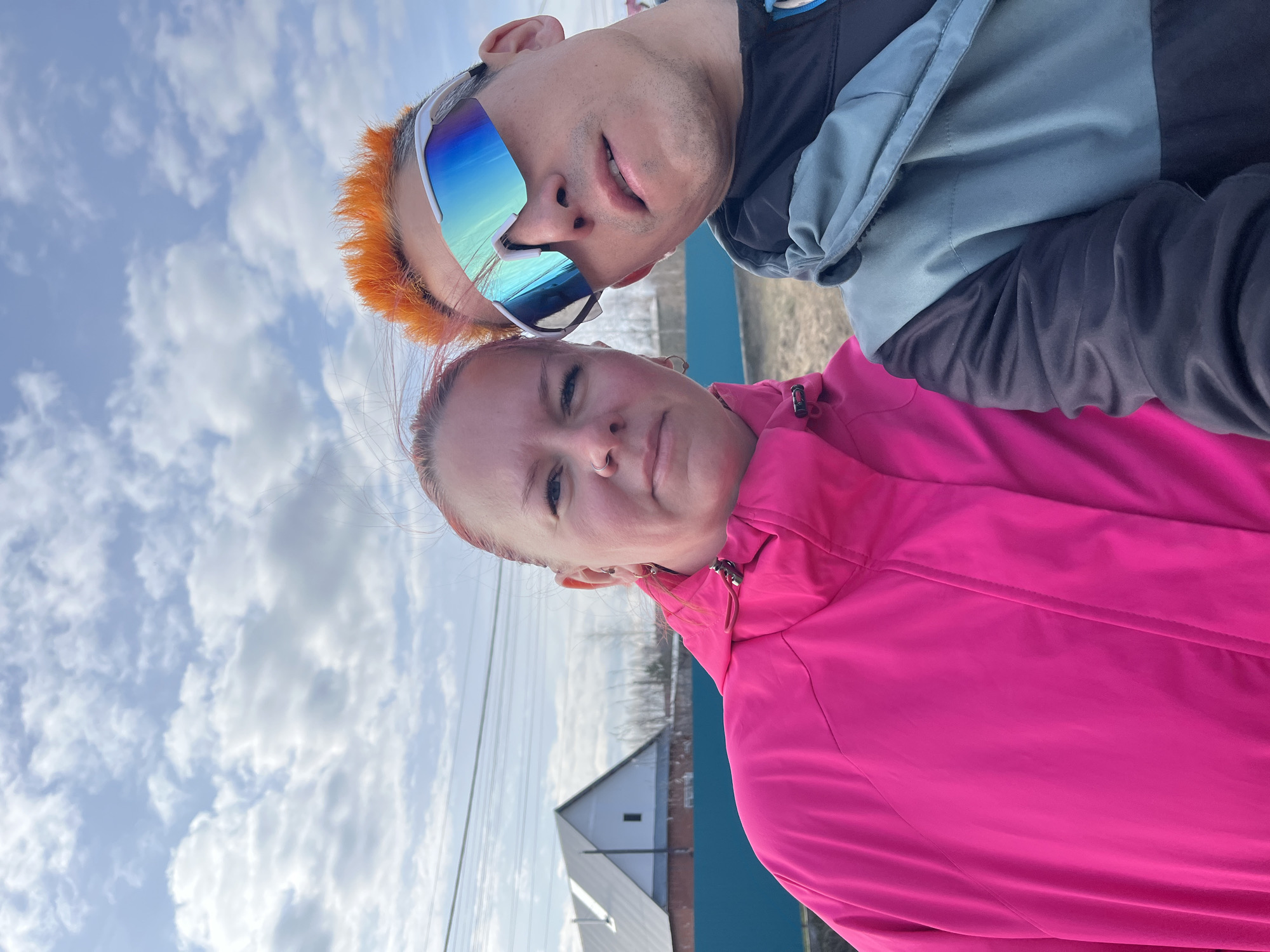 This vertical outdoor selfie captures two individuals, a man and a woman, against a bright, partly cloudy sky. The man is positioned above and slightly behind the woman, sporting vibrant orange-red hair and large, reflective sunglasses with blue and purple tints. He wears a dark jacket with light blue sections and has a neutral expression looking towards the camera.
The woman, in the foreground, wears a bright fuchsia jacket. Her hair, possibly dyed reddish, is styled back, and she has a nose piercing and earrings. She also looks towards the camera with a neutral or slightly serious expression.
The background reveals a daytime scene with scattered white clouds across a blue sky. Power lines traverse the upper background. Below them, a white-sided building with a dark roof, resembling a house or barn, is partially visible. A prominent, solid blue wall or structure spans the middle ground behind the woman. The overall setting appears to be an open outdoor space, likely rural or suburban in Chaikovsky, Russia, on an overcast but bright day. No visible text is present in the image.