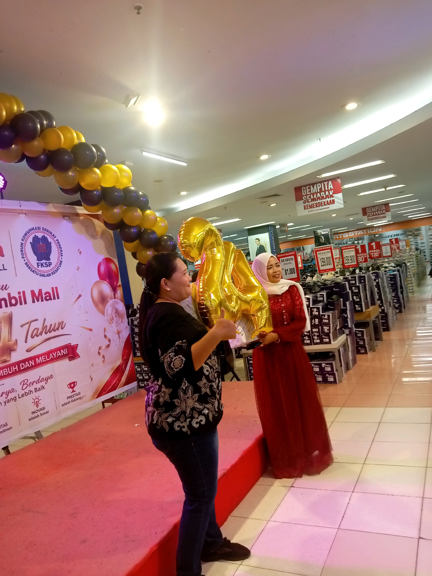 The image depicts an indoor scene, likely a retail environment, possibly a mall, given the presence of a banner that reads "Umbil Mall". The banner also indicates an anniversary celebration, stating "14 Tahun". Two women are the central figures. The woman on the left is holding a large gold balloon shaped like the number "4". She is wearing a black top with intricate white embroidery and dark blue jeans. The woman on the right is wearing a long, flowing red dress and a white hijab. She is smiling and appears to be interacting with the woman on the left, possibly holding a part of the same balloon. The background shows aisles of merchandise, stacked boxes, and retail signage, including price tags and advertisements. The flooring is tiled. Hanging from the ceiling are banners with Indonesian text, one of which appears to say "GEMPITA SEMARAK KEMERDEKAAN". Balloons, in shades of gold, yellow, and dark purple, are arranged in an arch above the banner and the women. The lighting suggests an indoor commercial space. While the specific location within Batam, Indonesia, cannot be definitively determined from the image alone, the presence of the "Umbil Mall" banner strongly suggests it is within that mall.