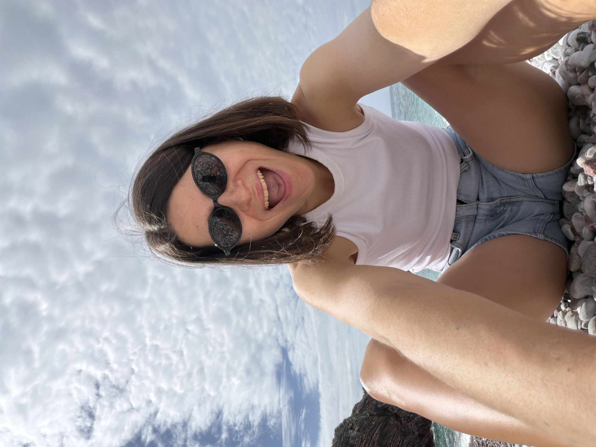 A young woman with short brown hair and sunglasses sits on a rocky surface, smiling at the camera. She is wearing a white tank top and denim shorts. The sky is visible in the background, featuring some scattered clouds. The photo was taken in Rafailovići, Montenegro.