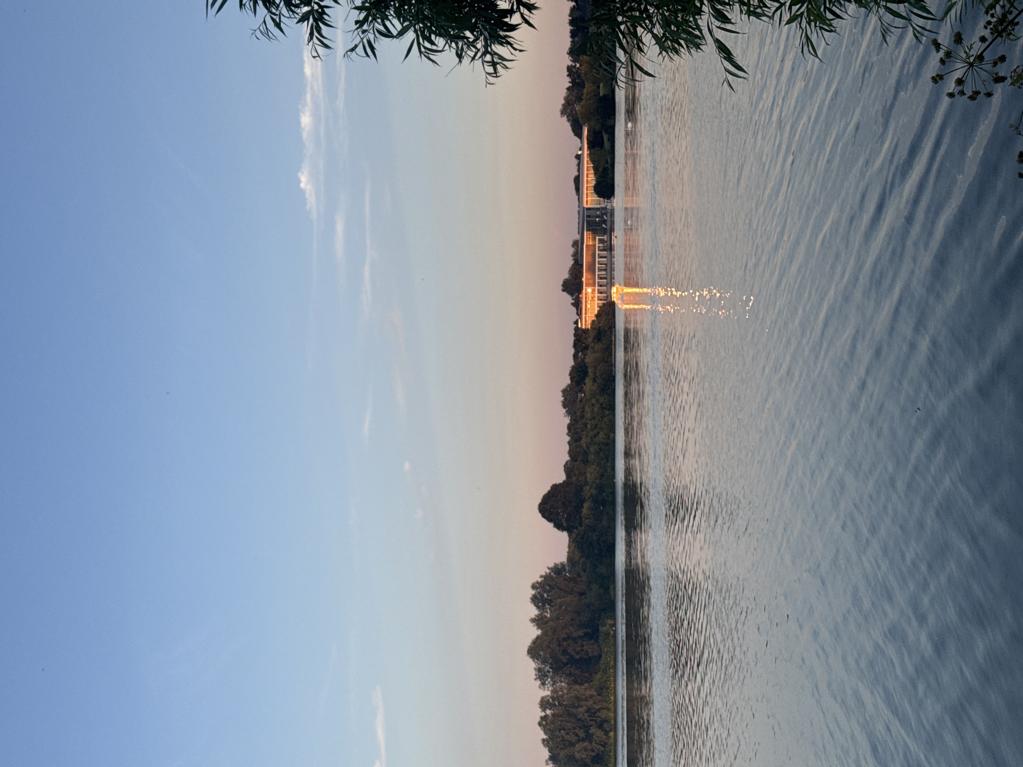 A wide shot captures a serene lake at sunset in Craigavon, United Kingdom. The sky is a gradient of pale blue fading to soft pink near the horizon. A long, modern building with large glass windows is visible across the lake, reflecting the warm, golden hues of the setting sun. The water's surface is calm, with gentle ripples creating shimmering reflections of the sky and the distant building. A lush line of trees forms a dark silhouette along the far bank. On the right side of the frame, the dark, leafy branches of a tree overhang the water, adding a touch of foreground detail. The overall atmosphere is peaceful and tranquil.