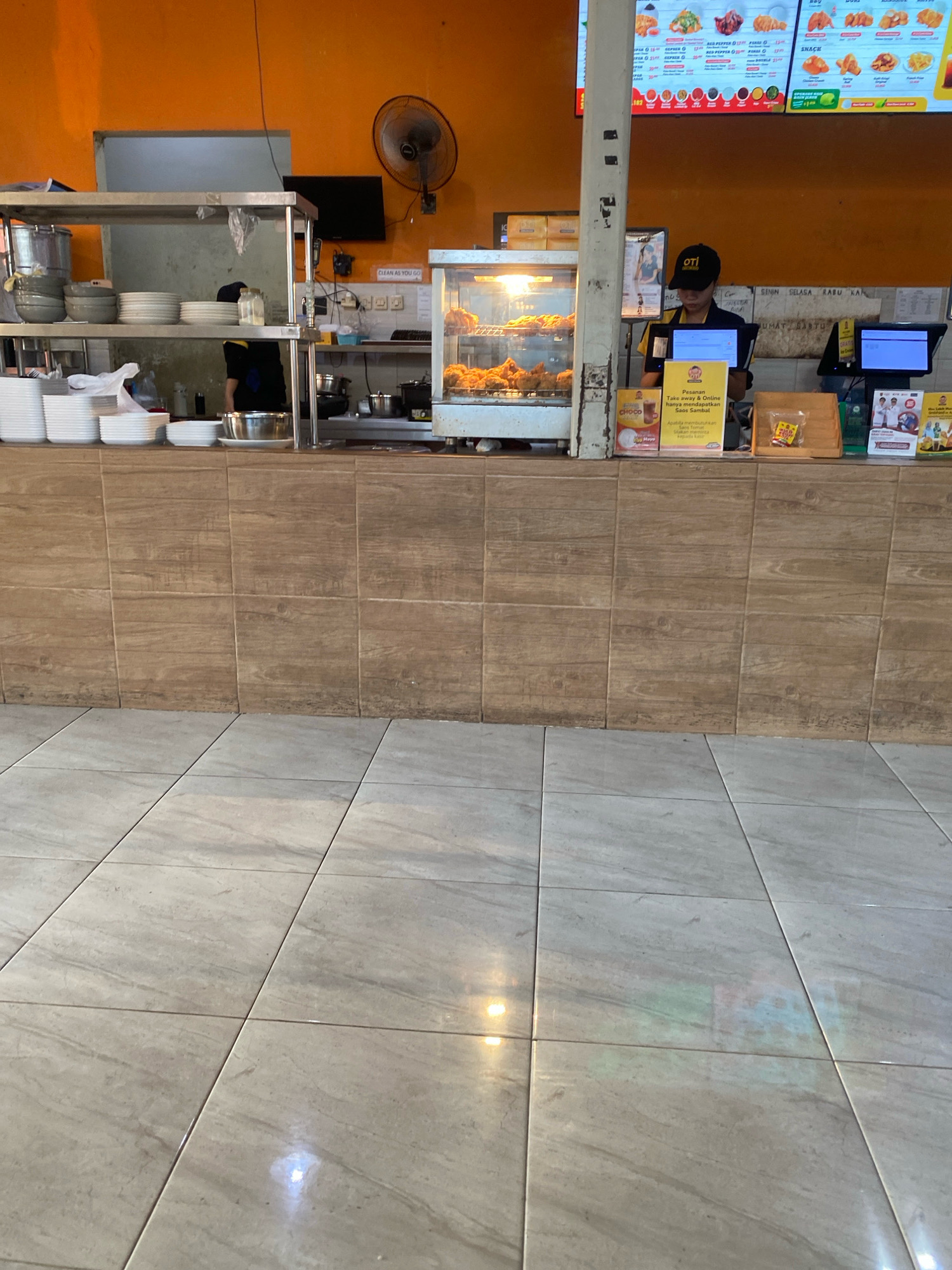 The image captures the interior of a fast-food restaurant, likely a fried chicken establishment, in Semarang, Indonesia. The scene is dominated by a service counter with tiled walls and a tiled floor.
In the background, an orange wall with a large menu board above the counter is visible, displaying various food items and prices. To the left of the counter, a stainless steel shelving unit holds stacks of plates and bowls, as well as some plastic containers and a large metal pot. A person wearing a dark uniform and a cap is visible behind the counter, interacting with a computer system. A glass display case filled with fried chicken is positioned on the counter.
On the counter itself, several items are arranged, including signs with information and possibly promotions, a small wooden dispenser, and what appear to be take-away containers. There are also computer monitors and a cash register. A standing fan is mounted on the wall behind the counter. The floor is tiled with a light-colored, marble-patterned tile. The overall impression is of a busy service area within a food establishment.
