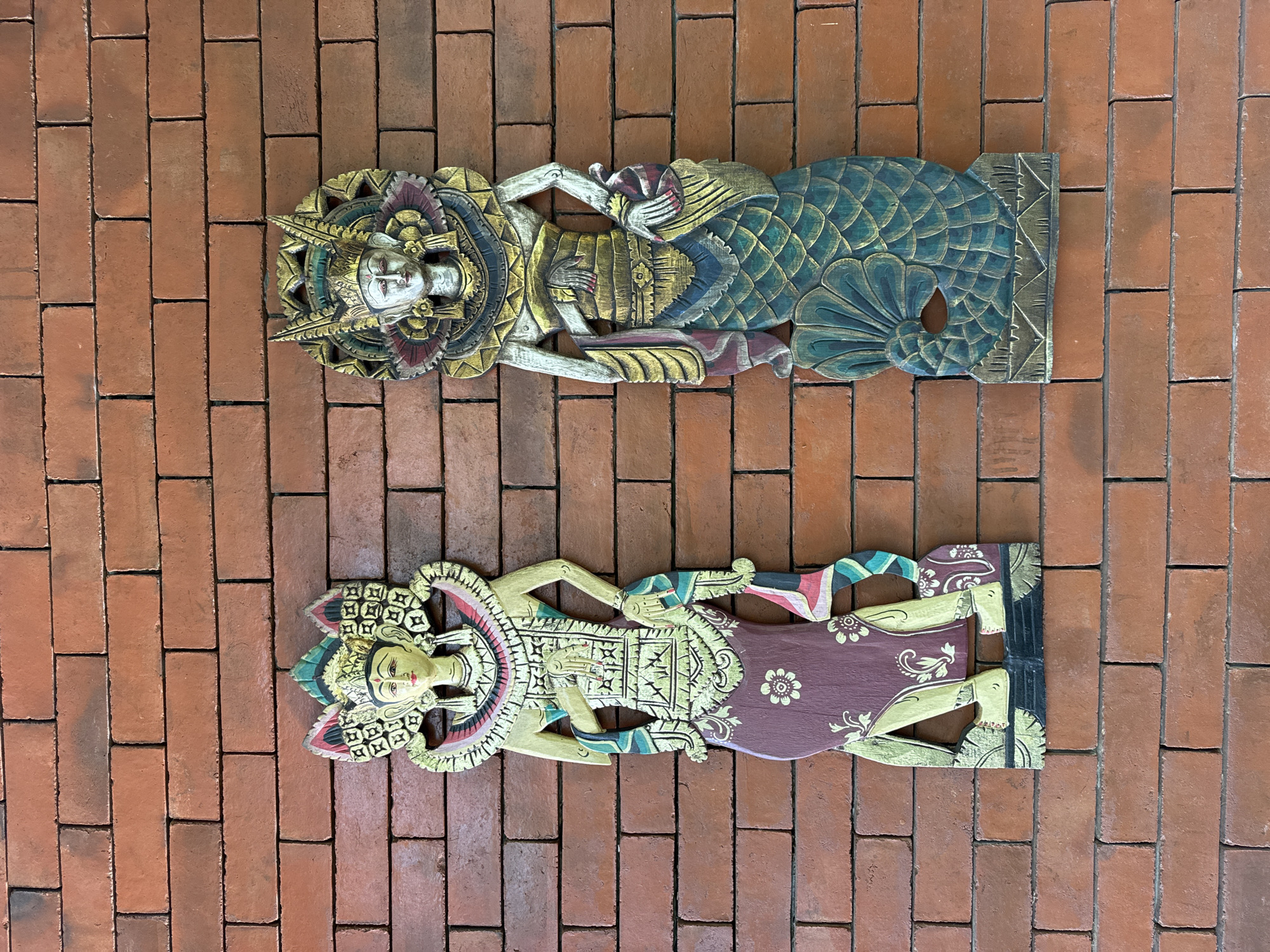 Two colorful wooden sculptures, depicting human figures, lie horizontally on a red brick surface. The top sculpture has a white face and is adorned with a golden headdress. It has a green, scale-like body with intricate golden detailing. The bottom sculpture features a similar golden headdress and detailed patterns on its body and clothing. The background consists entirely of red brick. This image was taken in the city of Rendang, Indonesia.