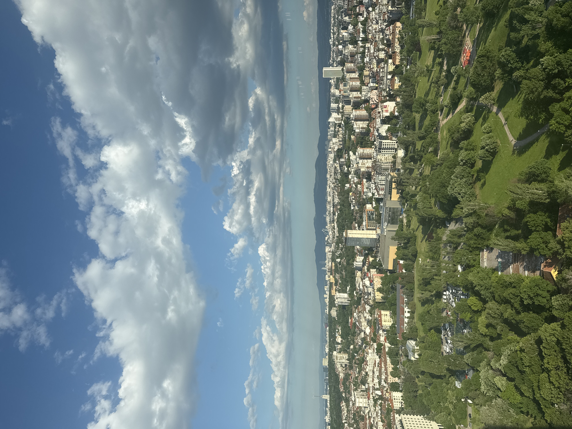 The image shows a view of Sofia, Bulgaria, possibly taken from a high vantage point. The left side of the image is dominated by a blue sky with numerous white clouds. The right side of the image features the urban landscape of Sofia, composed of buildings, green areas, and parks. A mountain range is visible in the distance in the center. The scene is well-lit, suggesting it is daytime. The urban area appears densely populated with a mix of residential and commercial structures.