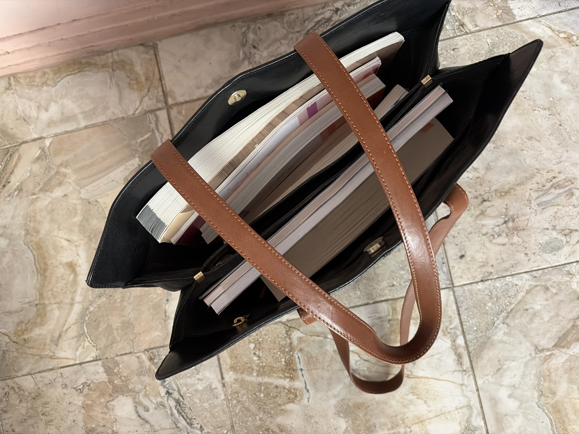 A black leather tote bag with brown leather straps is placed on a marble tiled floor. The bag is open and contains several books and magazines, some of which are stacked neatly. The flooring is light-colored marble with brown veining. In the background, a portion of a pink wall is visible. The image is taken from a high angle, looking down into the bag. There are no people or animals in the image. The scene is indoors. The location in the city of Burgas, Bulgaria is not determinable from the image.