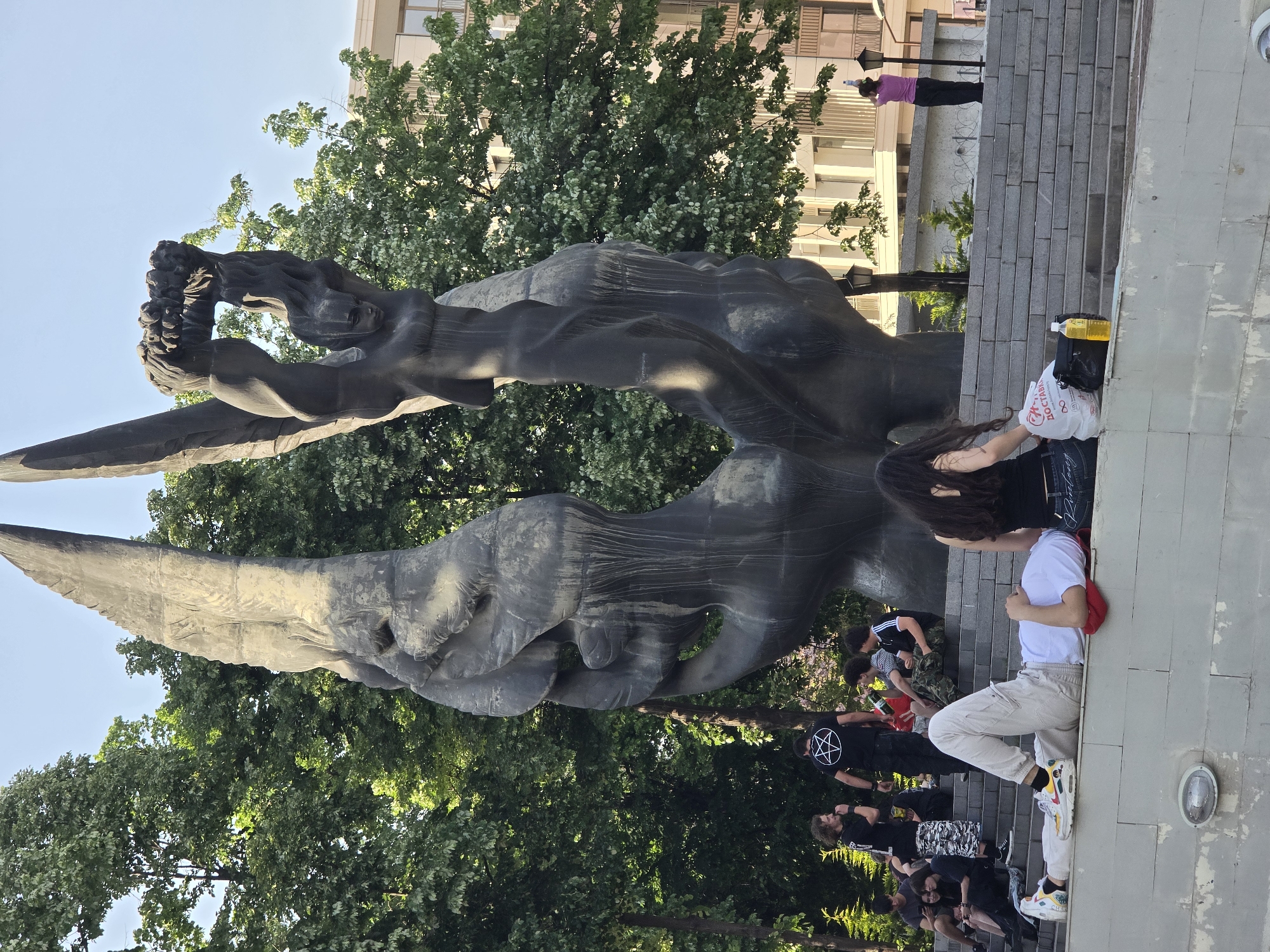 A bronze sculpture of a human figure with wings is set against a backdrop of green trees and a building with multiple windows. People are gathered at the base of the sculpture, some sitting on steps. One person in a white shirt and beige pants sits on a step with their legs outstretched. Another person with long, dark hair is seated nearby. A black t-shirt with a pentagram symbol is visible among the crowd. A yellow liquid in a bottle and a plastic shopping bag are placed on the steps near the people. The statue is the Revived Armenia monument in Yerevan, Armenia.