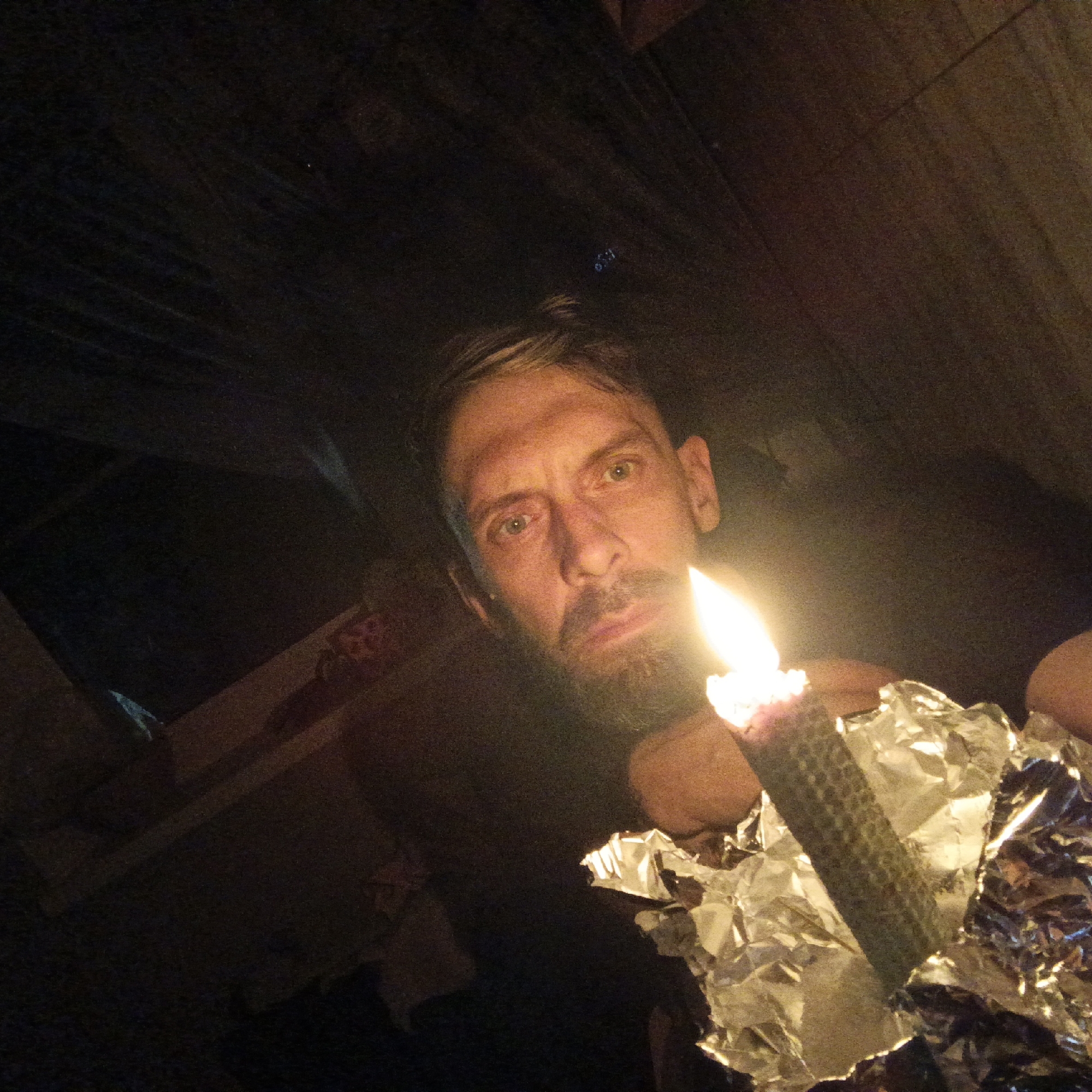 A close-up shot captures a man indoors, likely in Марковский, Russia, holding a lit candle wrapped in aluminum foil. The man has short, light brown hair and a goatee. The surroundings are dimly lit, with what appears to be shelving or a wall visible in the background. The candle's flame is the main light source, casting a glow on the man's face and the foil.