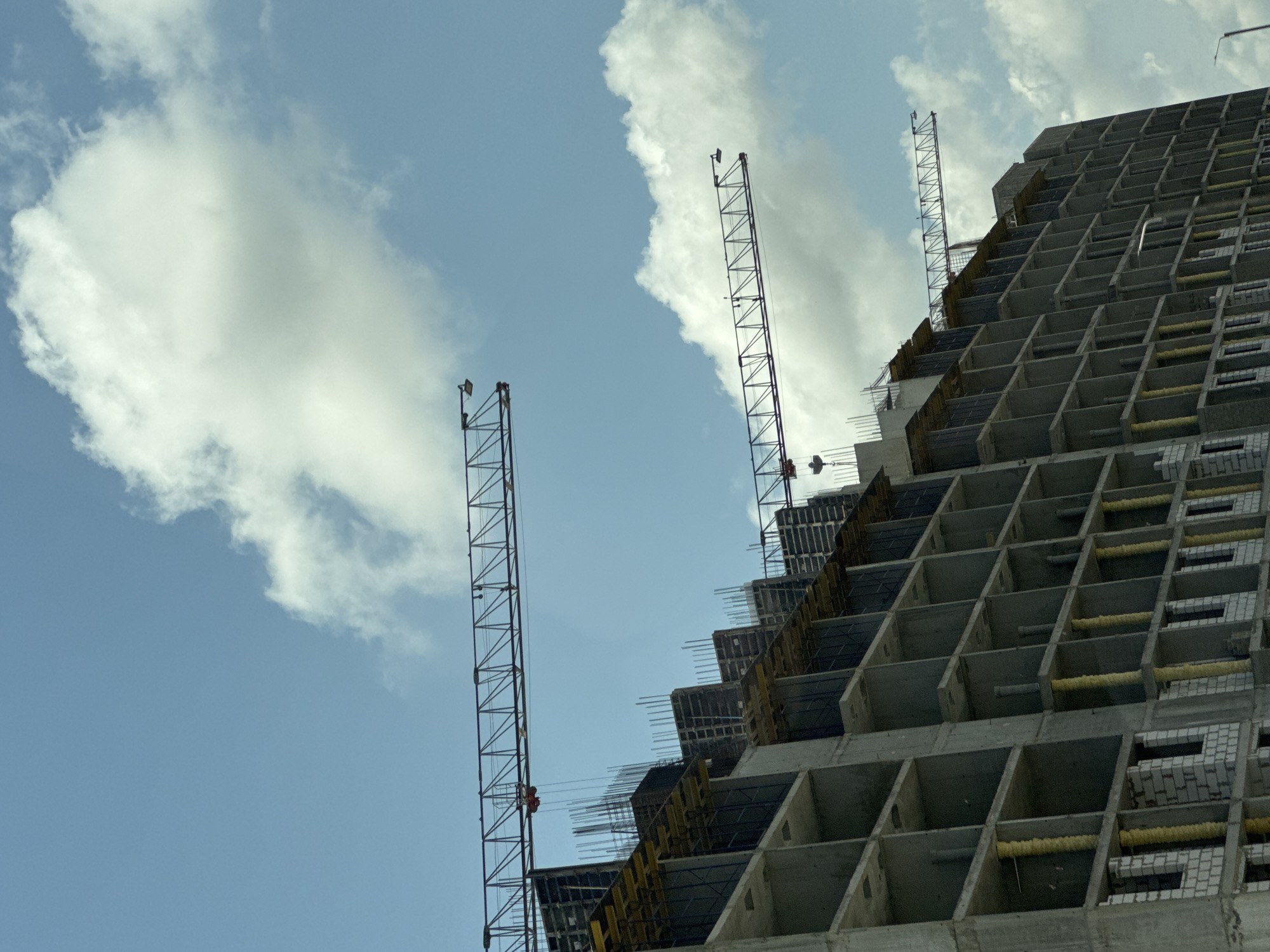 The image shows a construction site in the city of Mikhaylovsk, Russia. A large, multi-story building is under construction, with visible concrete floors, pillars, and walls. Several tower cranes are positioned around and above the building. The sky is blue with scattered white clouds. The lower levels of the building show some brickwork and exposed rebar. The scene appears to be captured from a low angle, looking up at the structure. There are no people visible in the image.