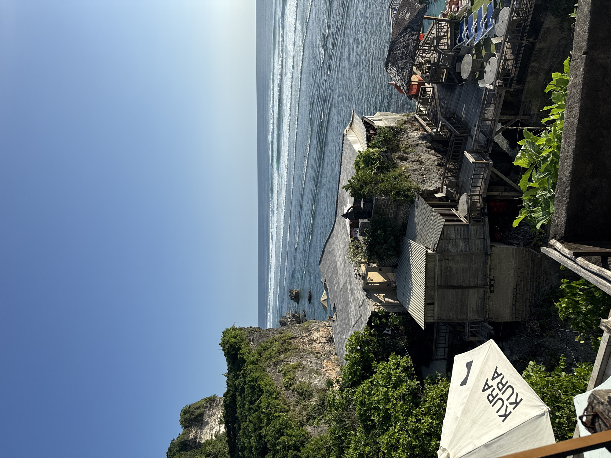 The image shows a seaside view in Kuta Selatan, Indonesia, capturing a mix of natural landscape and built structures. In the foreground and middle ground, there are buildings and cliffs covered with green vegetation, leading down to the ocean. The ocean stretches to the horizon, where it meets a clear blue sky. The buildings appear to be perched on the edge of the cliff, possibly part of a resort or restaurant complex. There are structures that may be wooden stairs connecting different levels, and lounge chairs are also visible. An umbrella is present in the foreground.