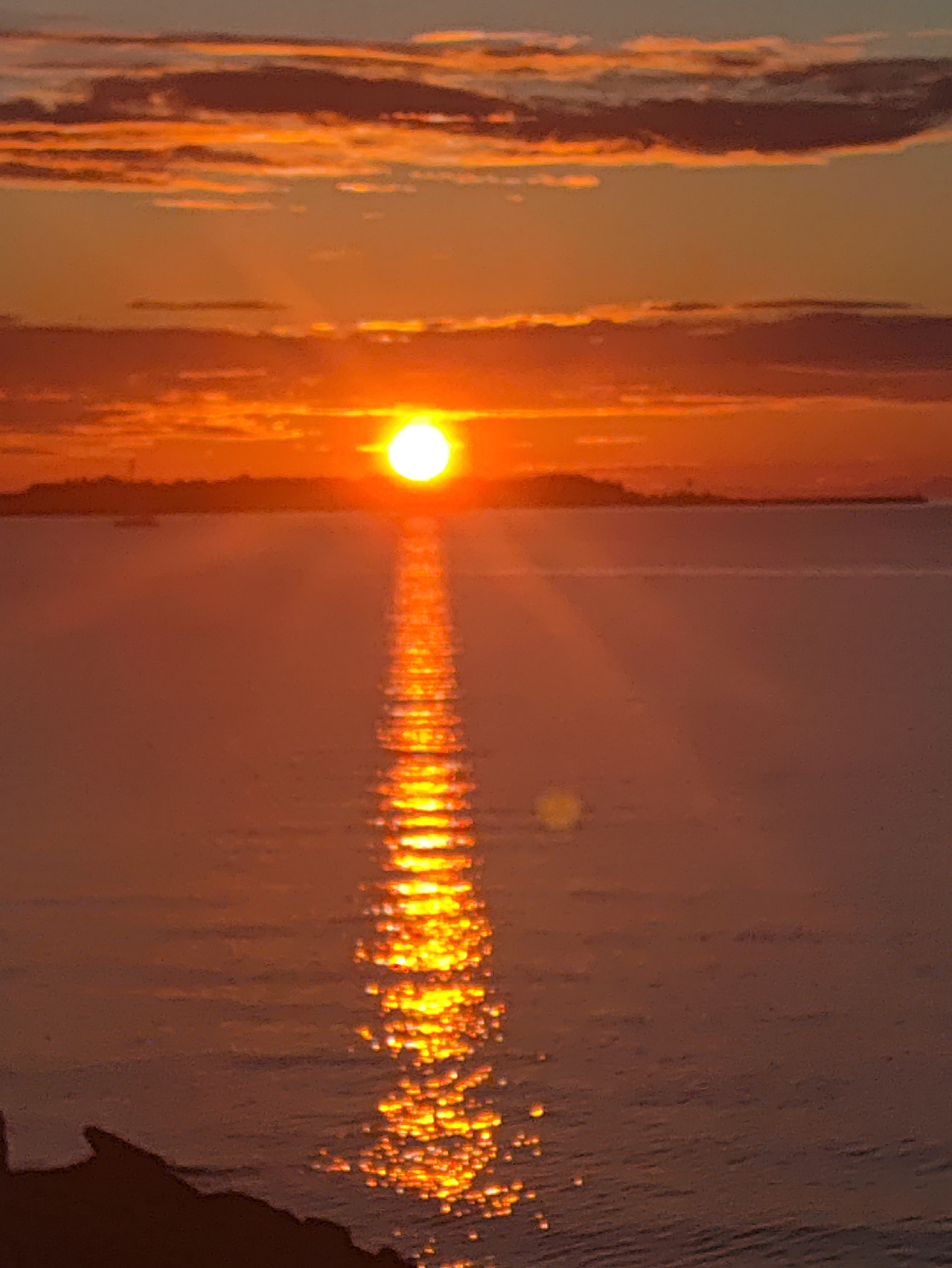 The image captures a sunset over the sea, with the sun appearing as a bright, glowing orb just above the horizon line. A reflection of the sun creates a shimmering, golden path across the water's surface, broken by ripples. The sky is a gradient of warm colors, transitioning from orange and pink hues near the horizon to softer blues and grays higher up, with scattered clouds tinged with the sunset's light. In the distance, a landmass, possibly an island or coastline, is silhouetted against the bright sky, with subtle structures or trees visible. The foreground shows a dark, indistinct shape, likely a rocky outcrop or shoreline. The overall scene conveys a sense of tranquility and natural beauty during twilight. This scene is not specifically identifiable as being within the city of Tallinn, Estonia, without further information or context.