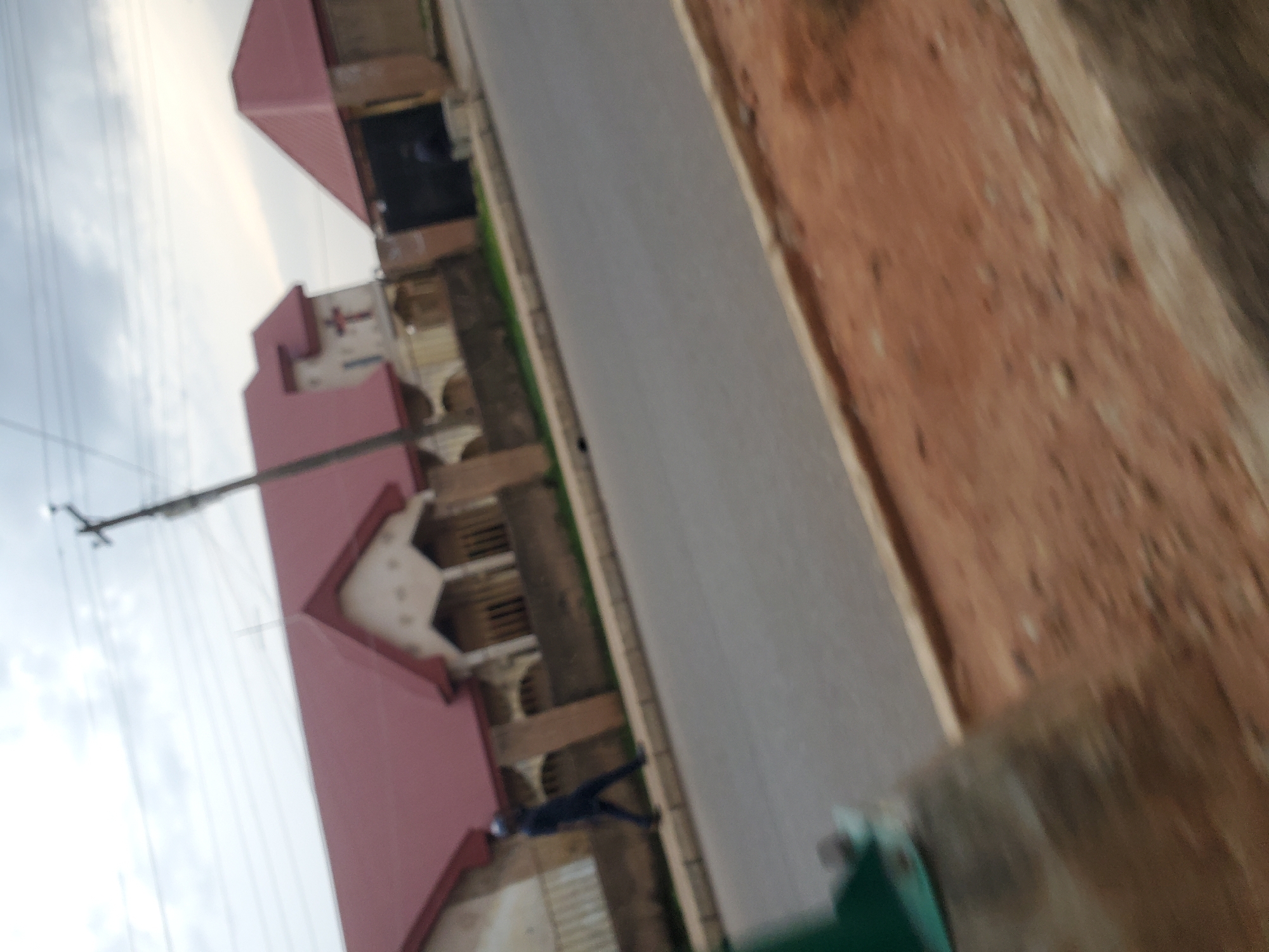 The image depicts a street scene in Bukuru, Nigeria, with a church prominently featured on the left. The church has a red, gabled roof and an architectural style that suggests it is a place of worship. A cross is visible atop one of the roof peaks. To the right of the church, another building with a similar red roof is visible, possibly an annex or a separate structure within the church complex. A utility pole with overhead wires stands near the church, indicating the presence of infrastructure.
In the foreground, a person in dark clothing is walking on the sidewalk adjacent to the road. The road itself appears to be paved with asphalt. The ground on the right side of the road is reddish-brown and appears to be unpaved or covered with dirt and gravel. The overall scene is somewhat blurry, suggesting the image was taken from a moving vehicle or with a camera that was not perfectly still. The sky is overcast with clouds.