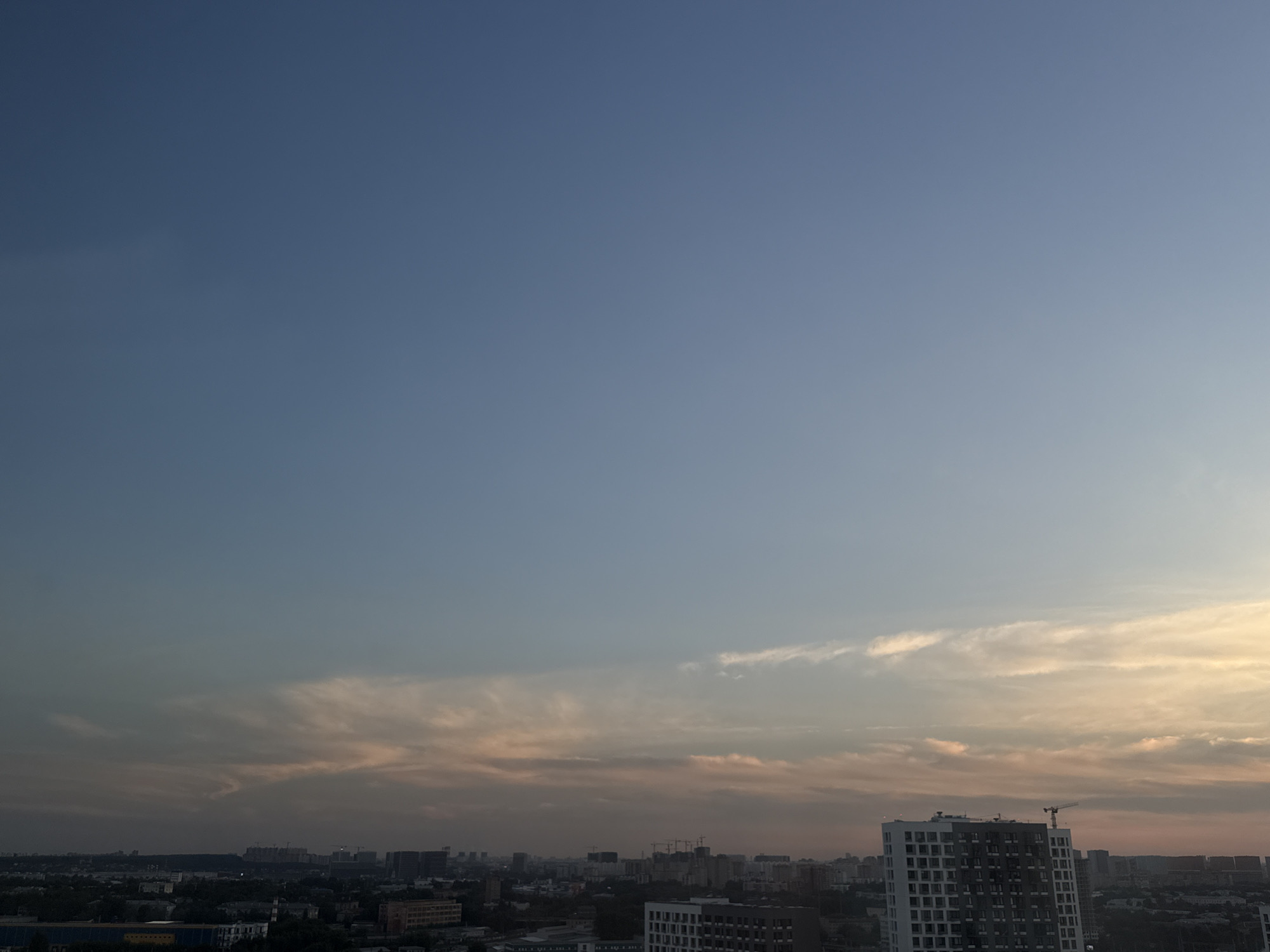 The image captures an aerial view of the city of Lyubertsy, Russia, at dusk. The sky dominates the upper portion of the frame, transitioning from a deep blue at the top to lighter shades of blue and pink near the horizon, suggesting the setting sun. Wispy clouds are scattered across the sky, catching the last rays of sunlight and casting soft, warm hues.
Below the sky, a sprawling cityscape unfolds. Numerous buildings, varying in height and architectural style, form a dense urban landscape. Some buildings appear to be residential, while others might be commercial or industrial. Construction cranes are visible in several areas, indicating ongoing development. In the foreground, a modern, tall residential building with a white and grey facade stands out. Further in the distance, the city stretches out, with more buildings and a visible expanse of trees and greenery interspersed within the urban fabric. The overall impression is one of a busy, developing city under a colorful evening sky.