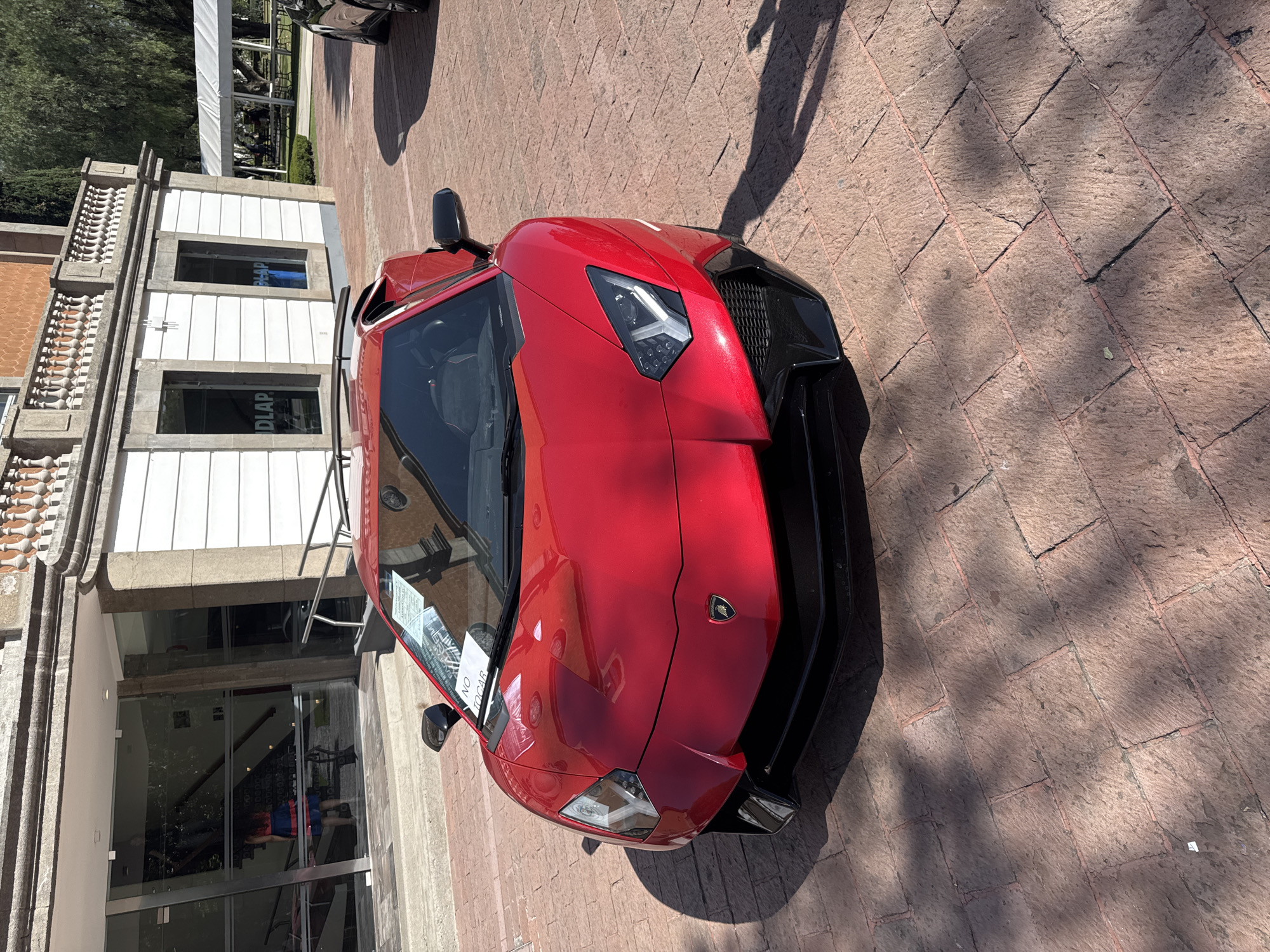 A vibrant metallic red Lamborghini sports car, likely a convertible, is the central subject of the image, parked on a paved reddish-brown ground resembling cobblestones or bricks. The car is positioned at an angle, with its front section prominent. Strong sunlight illuminates the scene, casting distinct shadows of a person and foliage onto the ground.
A white sign with red lettering, including the Spanish phrase "NO TOCAR" (Do Not Touch), is visible on the car's windshield. The Lamborghini emblem is clearly visible on the front hood of the vehicle.
To the left of the car stands a building with a blend of architectural styles. It features a decorative stone balustrade along an upper level, indicative of a classical design. The lower section of the building has modern white paneling and large glass entrances. Reflected or printed text, appearing as "DLAP" in reverse on the glass, is visible on the building's windows. This text, combined with the provided location context of San Andrés Cholula, Mexico, suggests proximity to the Universidad de las Américas Puebla (UDLAP).
In the upper background, lush green trees and a white tent structure can be seen, along with a portion of another dark-colored vehicle. The bright, clear lighting and sharp shadows indicate a sunny day, likely during the mid-morning or afternoon.