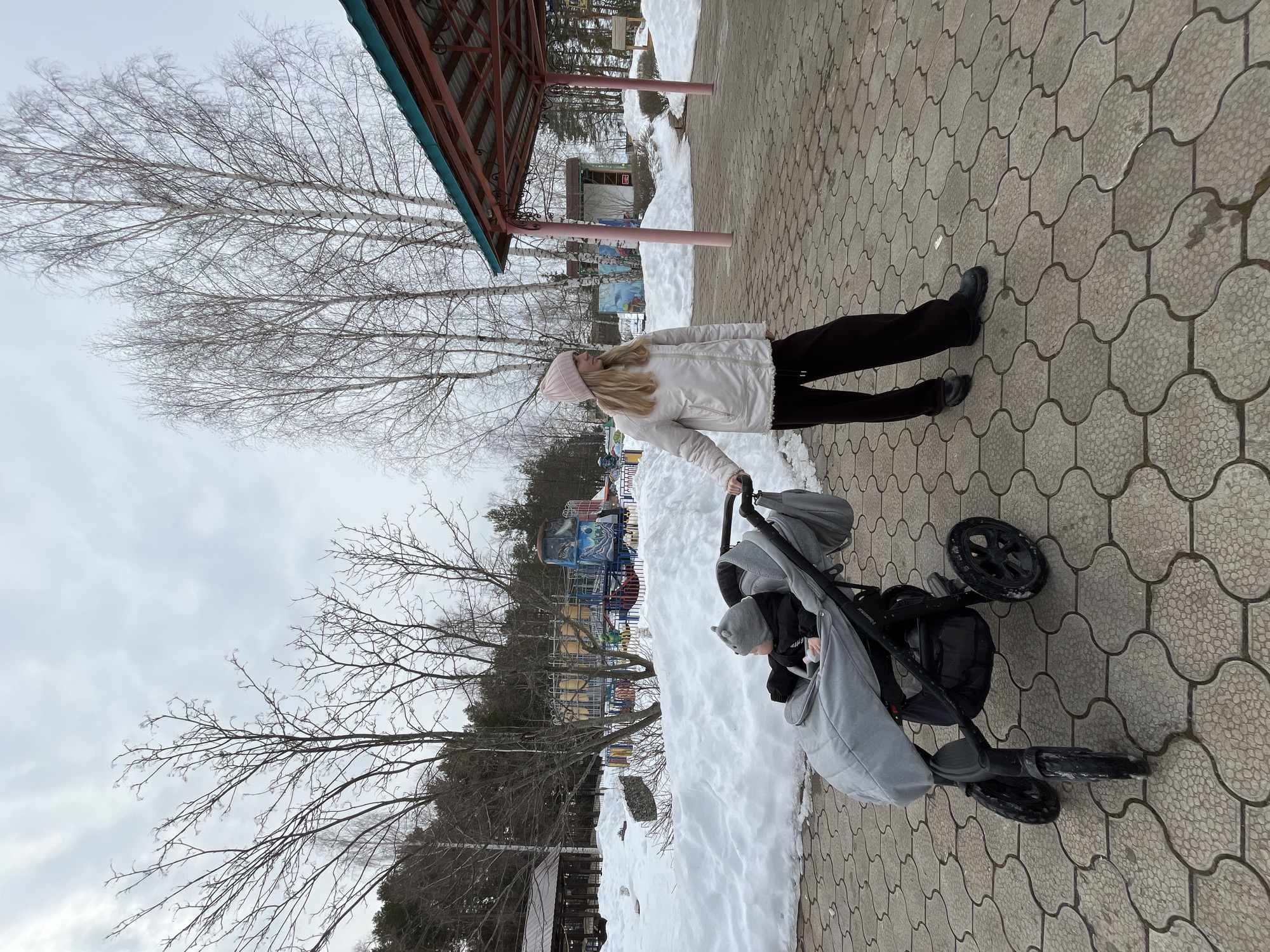 An adult female and an infant in a stroller are observed on a paved pathway in a park or recreational area within Chaikovsky, Russia. The pathway consists of light-colored, interlocking, textured hexagonal tiles. The environment indicates late winter or early spring, with significant snow cover visible in piles alongside the pathway and surrounding vegetation.
The female, appearing to be in her 20s, with long blonde hair, wears a light pink beanie, a cream-colored puffer jacket, dark trousers, and dark shoes. She stands to the right of a grey baby stroller with a black frame, holding its handle with her right hand, and is looking towards the left side of the frame. The infant, wearing a grey hat with ear-like details and dark clothing, is seated in the stroller, also looking towards the left.
In the background, leafless deciduous trees, including several birches, are prominent. A dense line of evergreen trees is visible further in the distance on the left. To the right, a structure with a dark teal sloped roof supported by pink pillars is partially visible. Elements of an amusement park are discernible in the mid-ground, beyond the snow and trees, including a yellow and blue carousel and a large blue and white vehicle-themed ride. The sky is overcast and light grey.