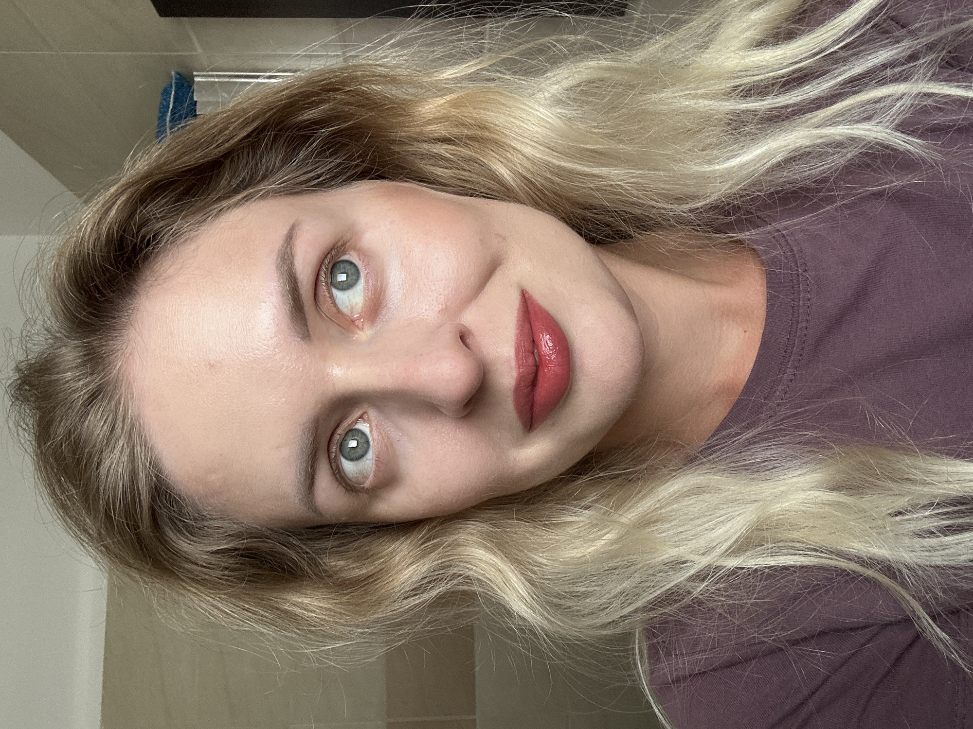 A medium shot shows a blonde woman with blue eyes and light skin. She is looking to her right with her lips slightly parted, revealing a glossy red lipstick. Her hair is wavy and falls around her shoulders. She is wearing a purple t-shirt. The background appears to be a bathroom with tiled walls. The scene is indoors, likely in a residential setting in Petah Tikva, Israel.