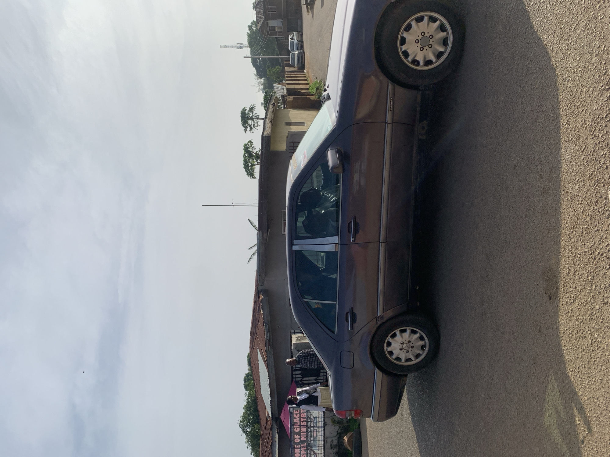 The image depicts a daytime scene on a paved road in Jos, Nigeria. A dark-colored sedan is parked or moving slowly, facing right, with its left side visible. The car has tinted windows and chrome accents. Inside the car, the driver is visible, and at least one passenger is also in the vehicle.
To the left of the car, a banner is displayed on a building. The banner has text in English, including "ZONE OF GRACE GOSPEL MINISTRY" and "SCHOOL OF THE SWORD". Two individuals stand near the banner; one is wearing a white top and a skirt, and the other is wearing a plaid shirt. A pink umbrella is also present near them.
Behind the banner and the people, a building with a corrugated metal roof is visible. Further back, more buildings, trees, and a telecommunications tower can be seen under a cloudy sky. Two other cars, a light-colored sedan and a silver hatchback, are parked further down the road in the background. The road surface is asphalt, and shadows are cast on it, indicating the sun's position.