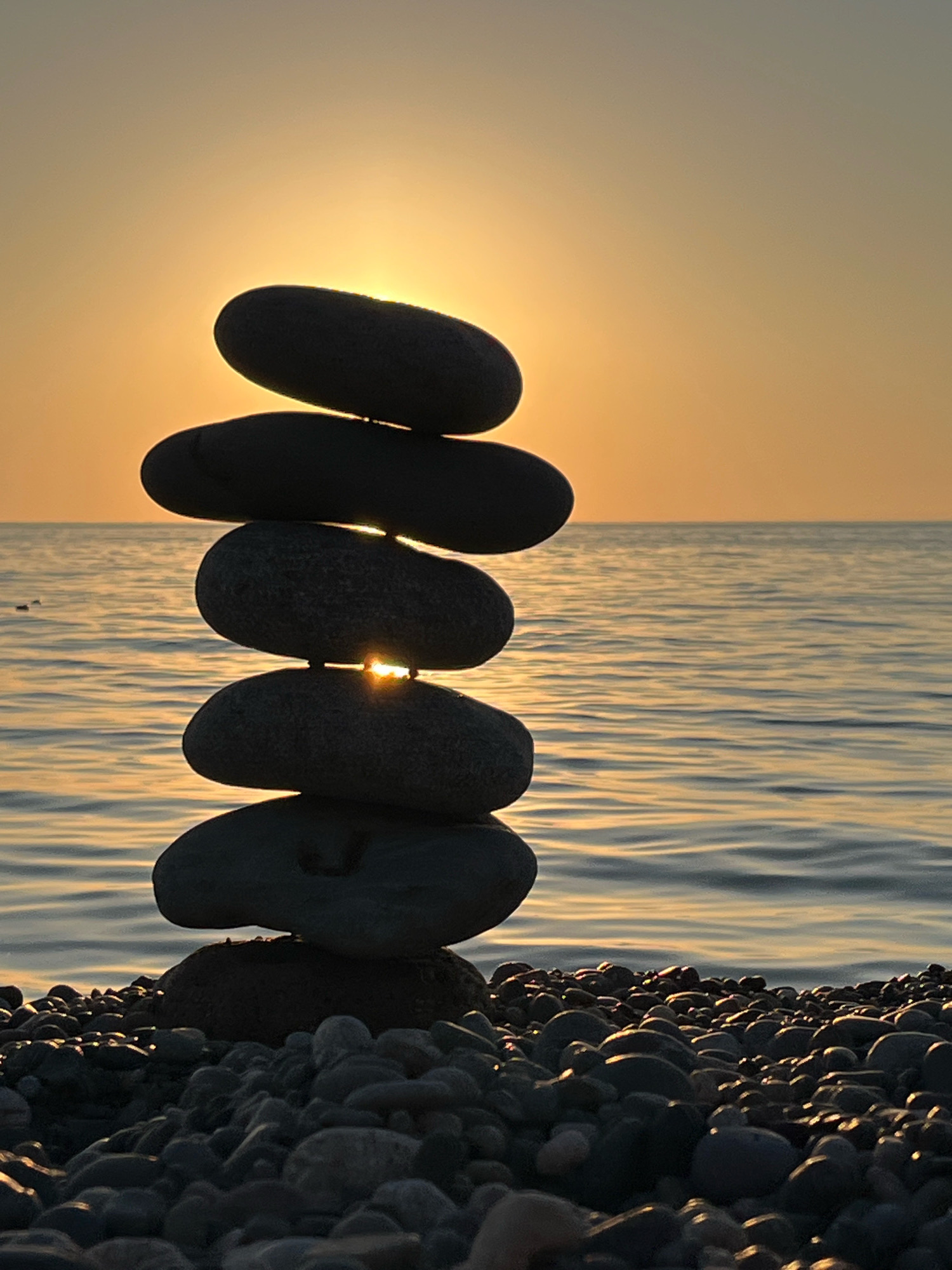 A close-up, full shot depicts a stack of six smooth, dark grey, oval-shaped stones balanced precariously on a pebble-covered beach. The stones are silhouetted against a warm, golden-orange sunset over a calm sea. The sun is peeking through the gaps between the stacked stones, creating shafts of light that illuminate the water. The water is a deep blue with gentle ripples reflecting the sky's warm hues. The beach in the foreground is covered in a dense layer of various sized grey pebbles, some catching the light. The scene is located in Kobuleti, Georgia.