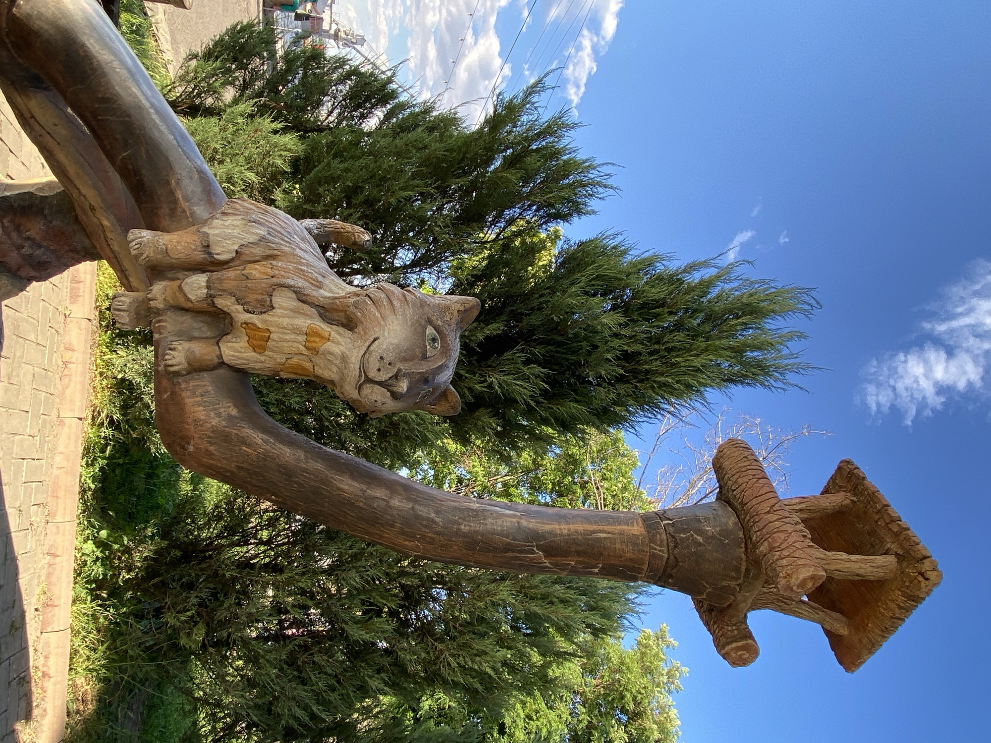 In an outdoor setting under a clear blue sky with scattered clouds, a wooden sculpture of a cat sits on a thick, curved wooden branch. The cat sculpture is light brown with darker markings resembling patches and has a friendly expression. Above and behind the cat, a larger wooden structure resembling a birdhouse or feeder, made of rough-hewn wood, is perched atop a thick, dark brown wooden pole. The pole is bent, with the cat sculpture positioned on its lower section. The background consists of lush green trees and foliage. The ground in the foreground is paved with light-colored bricks. The overall scene suggests a whimsical outdoor art installation in an urban park or public space.
