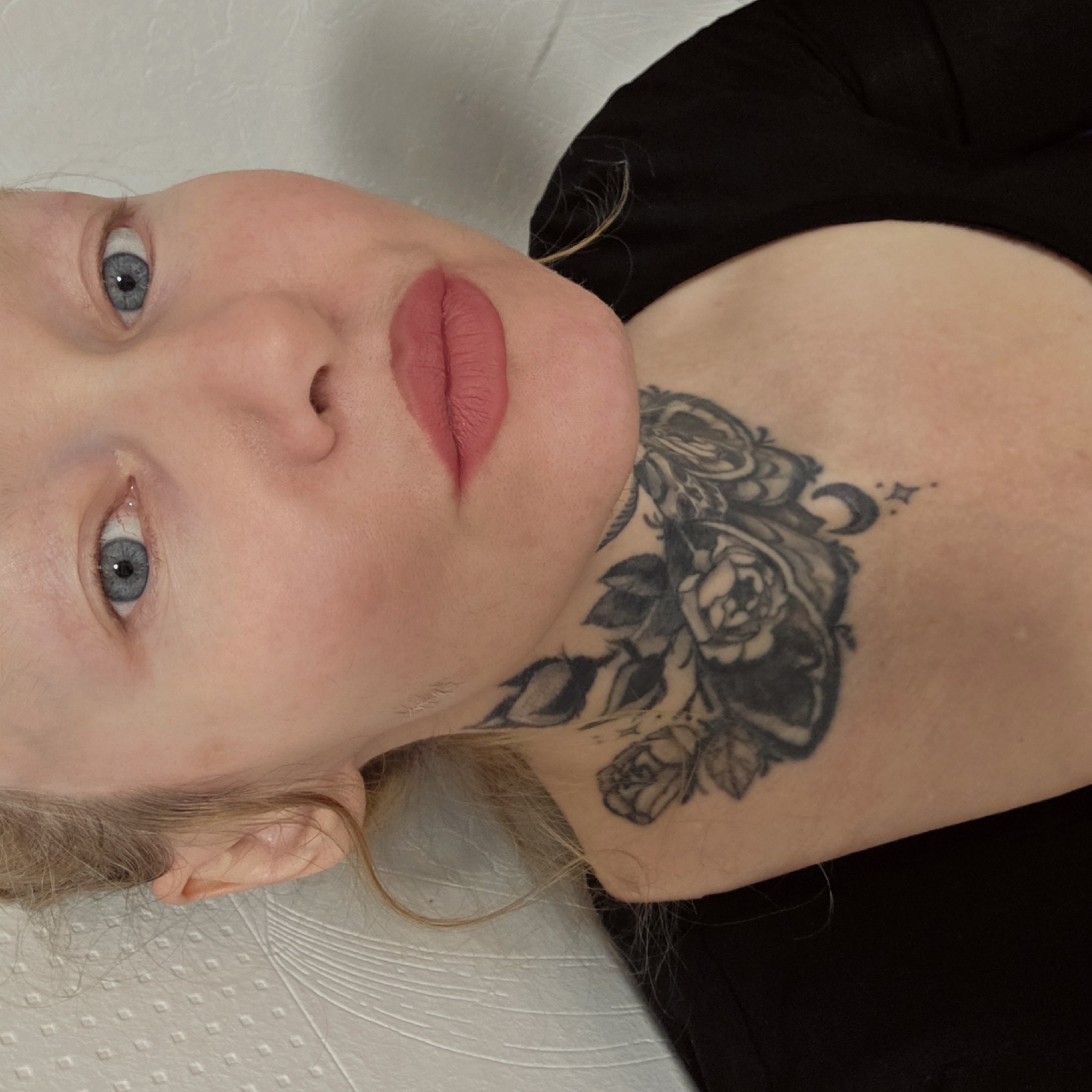 A pale-skinned person with blue eyes and blonde hair is lying down, facing the camera. They have pink lipstick on. A black tattoo featuring roses, leaves, a crescent moon, and stars is visible on their neck. The person is wearing a black top. The background is a white textured surface. There is no information in the image to suggest the location is Saint Petersburg, Russia.