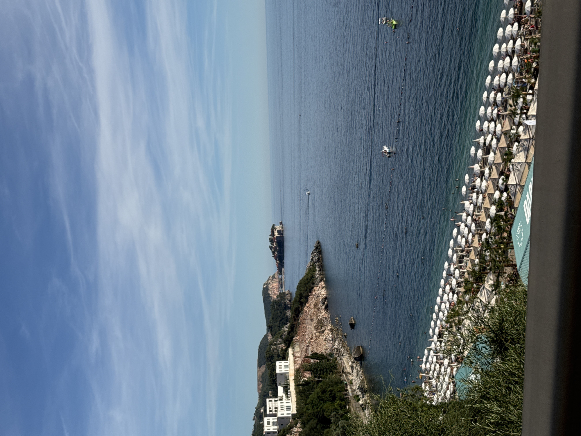 The image presents a wide, elevated view of a sun-drenched coastline, identified as Kamenovo, Montenegro. The scene is dominated by the vast expanse of the deep blue Adriatic Sea meeting a bright sky adorned with wispy white clouds.
In the foreground, a densely packed beach stretches along the shoreline. Numerous white umbrellas and some beige cabanas are arranged in neat rows, casting small shadows on the sand. People are visible beneath the umbrellas, some reclining and others appearing to be walking. The water at the edge of the beach is a clear turquoise, transitioning to a darker blue further out. Several individuals are in the water, swimming or wading. A thin, dark rope demarcates an area in the sea, likely for swimmers.
To the left, a rocky, tree-covered coastline juts into the sea. White, modern buildings with clean architectural lines are perched on the hillside, overlooking the water. Below these buildings, a path is visible, winding along the coast. Further out to sea, a small, fortified island with buildings is situated off the mainland.
Several boats are scattered across the water. One white boat is near the middle of the frame, and another, larger vessel is closer to the shore, on the right side of the image. A yellow inflatable watercraft is also visible on the far right. The overall impression is one of a vibrant coastal resort on a clear, pleasant day.