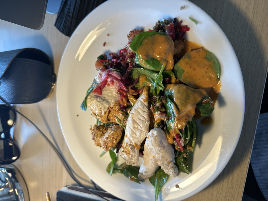 A white oval plate containing a prepared salad is centrally positioned on a light-colored wooden desk surface in the City of London, United Kingdom. The salad comprises grilled white meat strips (likely chicken), several breaded or crispy fried pieces (possibly falafel or seasoned chicken), fresh green spinach leaves, and a grain mixture containing visible red and darker fragments (likely quinoa and diced vegetables). A pale, creamy sauce and a reddish liquid are present on some of the crispy pieces, while a vibrant orange-red dressing is liberally applied across the entire salad. In the background, partially visible items on the desk include dark sunglasses, a black cable, a dark blue or black leather accessory, a corner of a grey electronic device, and a stack of black, thin objects. No individuals are depicted.