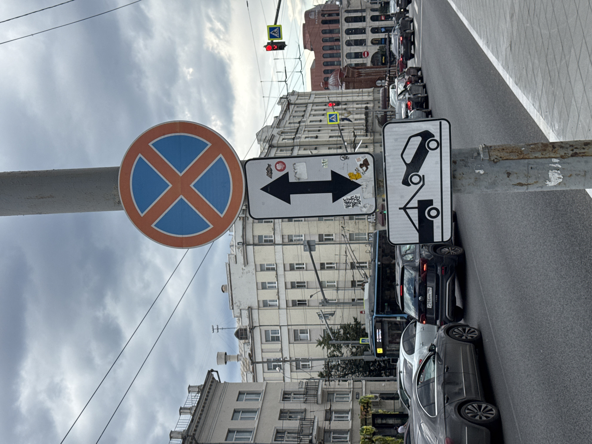 This image shows a street scene in Yekaterinburg, Russia, on a cloudy day. The foreground features a metal pole with several traffic signs. The topmost sign is a circular red and blue sign with an orange "X" shape, indicating no stopping. Below it, a rectangular sign displays a black double-headed arrow, signifying two-way traffic. Further down, another rectangular sign illustrates a tow truck hooking up a car, warning of towing.
In the background, several cars and a blue bus are on the road. The architecture suggests an urban environment, with multi-story buildings lining the street. Traffic lights and street lamps are visible. The road surface is asphalt, and a pedestrian sidewalk is present on the right side of the image. The overall scene is typical of a busy urban street.