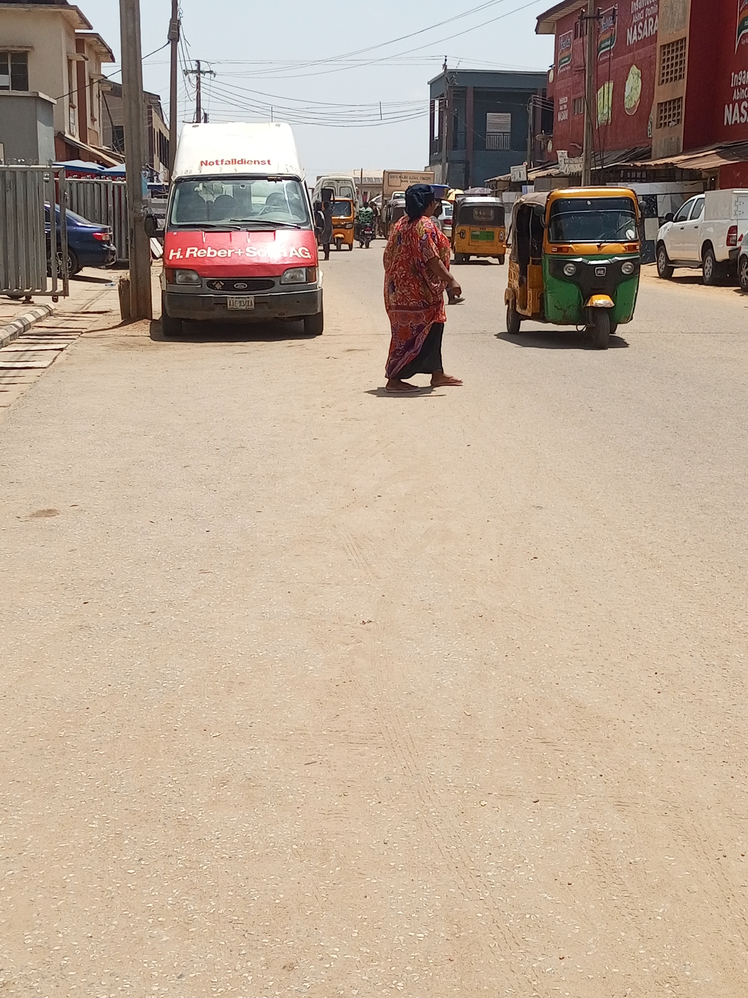 The image shows a street in Maiburiji, Nigeria. A woman in a patterned orange and black dress is walking across the street. A yellow and green auto rickshaw is driving on the right. A white van with "Notfalldienst" and "H. Reber+Sohn AG" written on it is parked on the left. Other auto rickshaws and vehicles are visible in the background. Buildings line the street. The sky is overcast. The street is paved and dusty.