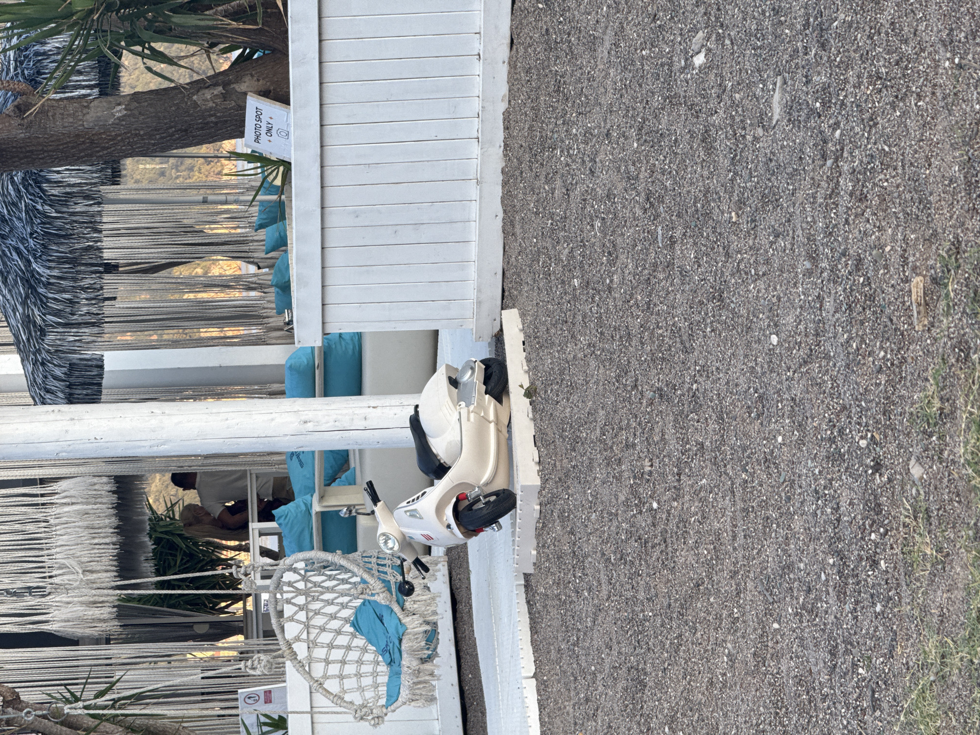The image depicts a scene at a beachside establishment in Boreti, Montenegro. In the foreground, a small, cream-colored scooter is parked on a white platform. The scooter has a black seat and red accents. Behind the scooter, a white wooden fence runs horizontally across the frame. To the left of the scooter, a white rope-strung hammock chair with a teal cushion is visible, hanging from a white structure. In the background, there are white macrame curtains, palm trees, and people standing near a seating area with teal cushions. A sign on a wooden post reads "PHOTO SPOT ONLY". The ground is covered in dark grey gravel.