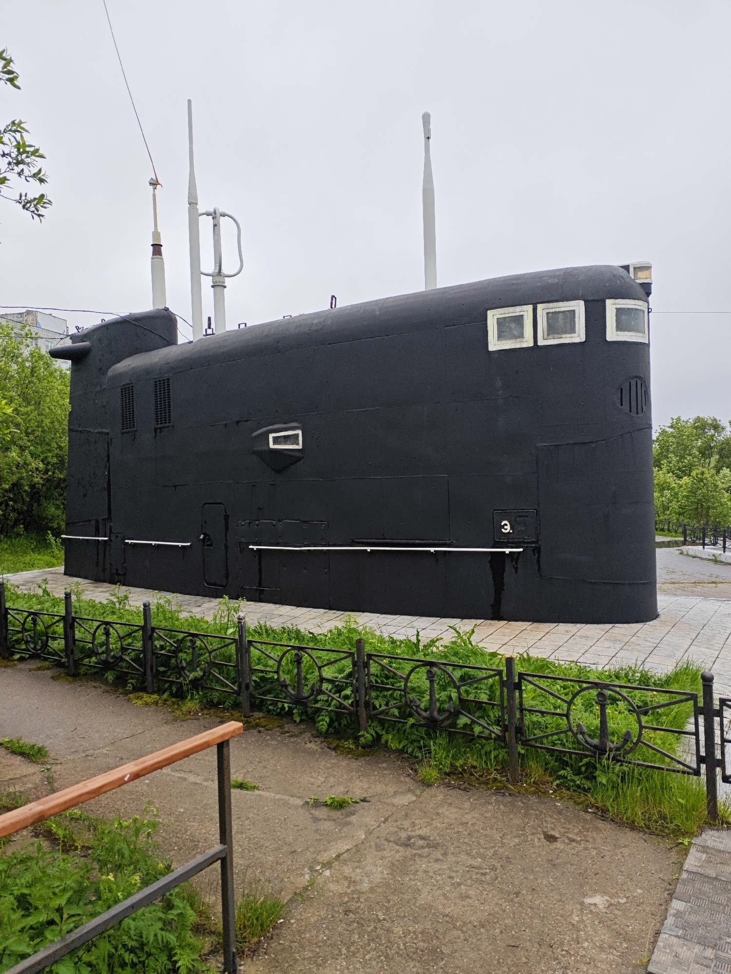The image shows a black, decommissioned submarine displayed outdoors in the city of Polyarny, Russia. The submarine is mounted on a concrete platform surrounded by a black metal fence with anchor motifs. Green grass and some vegetation grow around the base of the submarine and behind the fence. A paved walkway is visible in the foreground, with a wooden handrail. The submarine has several rectangular windows and visible antennas on its upper section. A number "3" is painted on the side of the submarine. The sky is overcast.