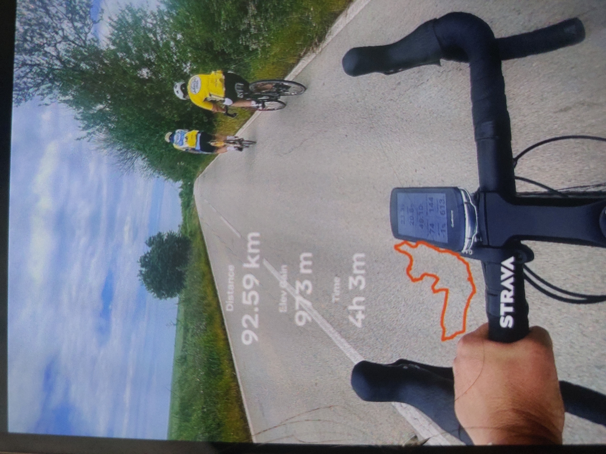 The image is a first-person perspective shot from a bicycle, showing a cyclist's handlebars, a Strava device displaying ride statistics (92.59 km distance, 973 m elevation gain, 4 hours 3 minutes time), and a GPS route outlined in red. In the background, two other cyclists are visible on a paved road bordered by green foliage and a blue sky with clouds. The display screen shows additional information: a speed of 20.8, time of 48.10, heart rate of 74/144 and elevation of 613. The GPS route is in a location outside of the city of Tsarevo, Bulgaria.