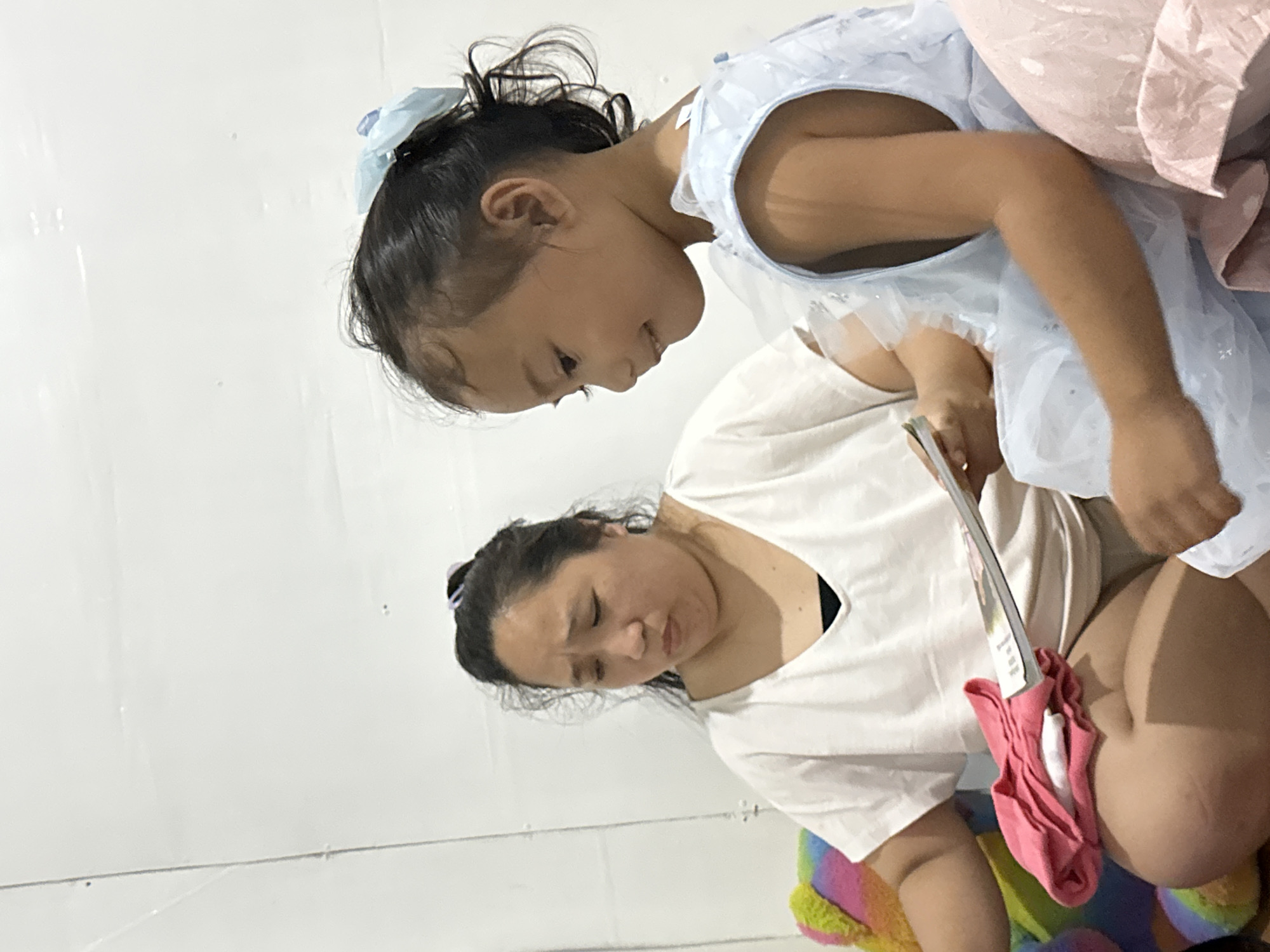 In a domestic indoor setting in Davao City, Philippines, a woman with dark hair tied back sits and holds a book. She is wearing a white t-shirt and has a pained expression on her face. Next to her, a young girl wearing a light blue dress with a bow in her hair smiles as she looks at the book. The background is a plain white wall.