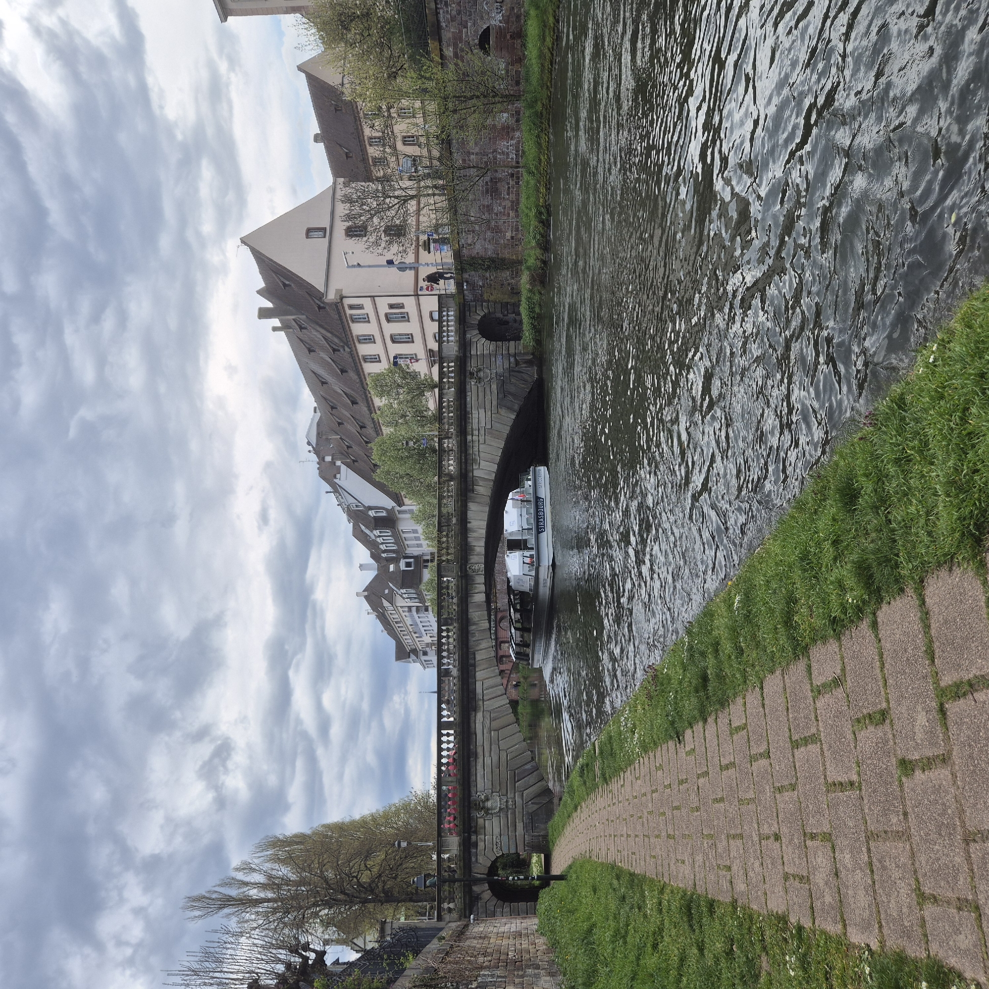This image shows a view of a canal in Strasbourg, France, with buildings along the banks. A tour boat labeled "Strasbourg" is passing under a stone bridge. A stone walkway runs alongside the canal, with grass growing between the stones. The sky is cloudy. Two people are visible standing on the bridge above the boat.