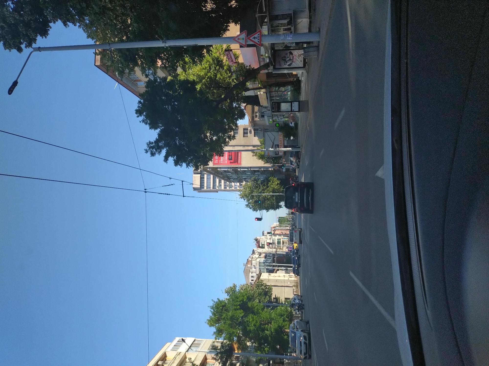 The image captures a view from a vehicle driving down a multi-lane street in Stara Zagora, Bulgaria. The street is lined with buildings of varying heights and architectural styles, some featuring balconies and air conditioning units. Tall trees with lush green foliage are interspersed along the sidewalks and between buildings. Power lines and streetlights are visible overhead. Traffic signals are present, with one red light visible in the distance. Several cars are driving on the road, including a silver hatchback, a black SUV, and a yellow sedan. A motorcyclist is also visible. On the right side of the street, there is a bus stop shelter with advertisements and a trash receptacle. Traffic signs, including a pedestrian crossing sign and a warning sign, are also visible. The sky is clear and blue, suggesting a sunny day. The overall scene depicts a typical urban environment in Stara Zagora.