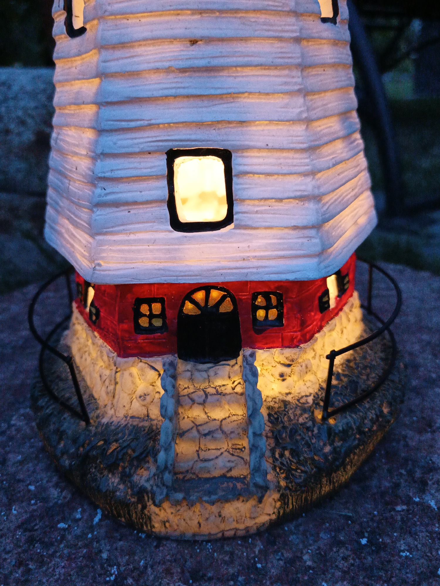 A close-up, low-angle shot captures the bottom half of a decorative, illuminated lighthouse at dusk. The lighthouse has a white, ribbed upper section with a single, glowing window. Below, a red section features two small, square windows with yellow panes and a black-arched doorway with a fanlight pattern that also glows yellow. The base of the lighthouse is textured to resemble stone and is illuminated from within. A stone pathway leads up to the doorway. A dark, curved railing surrounds the base. The lighthouse sits on a dark, pebbly surface, possibly a pathway or ground. The background is dark and blurred, suggesting foliage or other out-of-focus elements. The scene does not depict any people, nor is there any indication of a specific location in Лосева, Belarus.