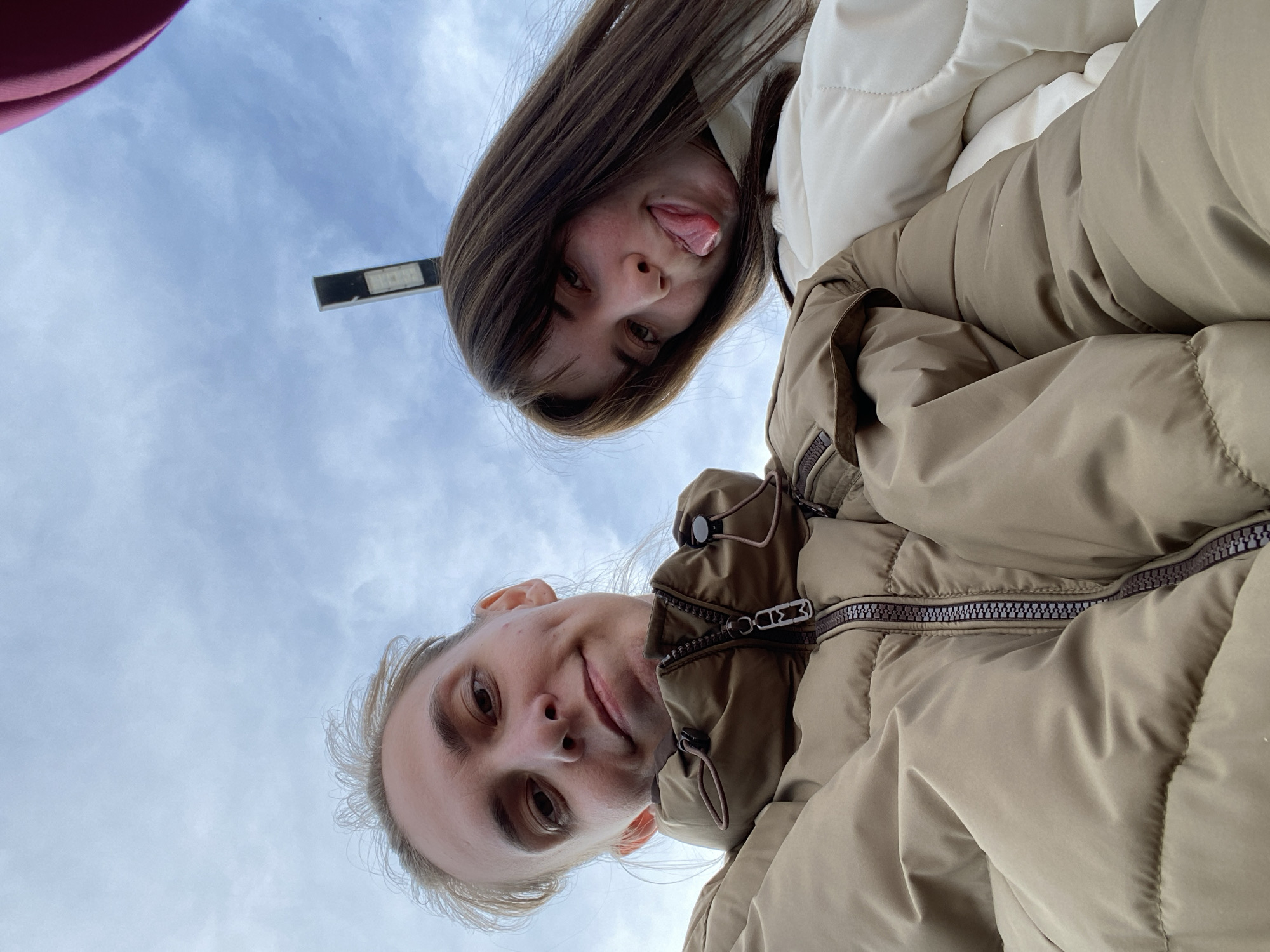 Two people are looking down at the camera in an outdoor setting. The background shows a cloudy sky. One person has brown hair and is sticking their tongue out. They are wearing a white puffy jacket. The other person has blonde hair and is wearing a tan puffy jacket with zippers. The location is assumed to be in the city of Chaikovsky, Russia. There is a light pole visible in the background.