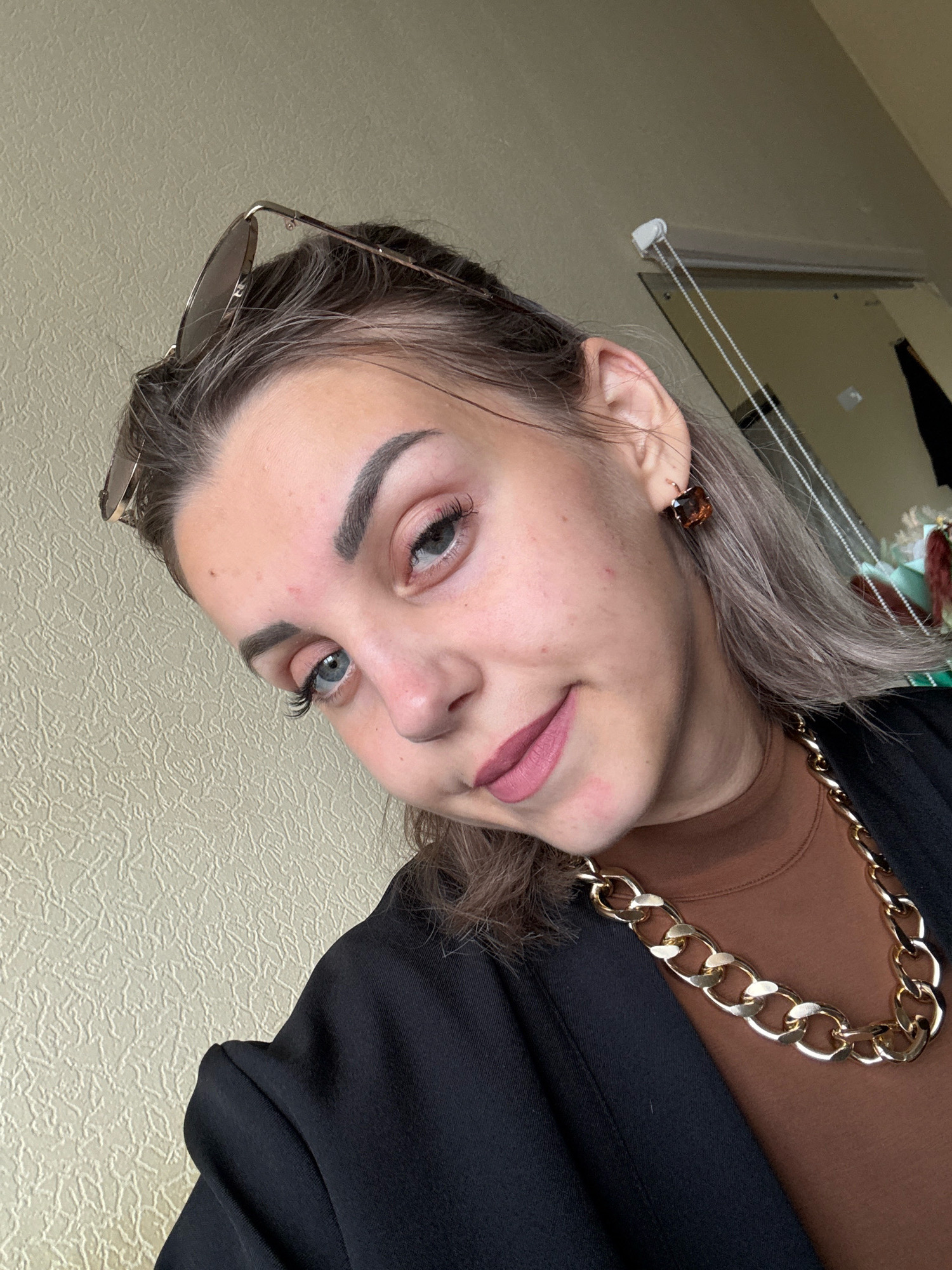 The image is a selfie of a young woman, likely taken indoors. The woman has light brown hair with some lighter streaks, styled down. She is wearing makeup, including mascara, eyeliner, and lipstick in a pinkish-mauve shade. She has prominent eyebrows and light blue eyes. She is wearing a dark blazer over a brown turtleneck and a thick gold chain necklace. She also has on square-shaped brown earrings. On top of her head, resting against her hair, are a pair of rose-gold colored sunglasses. The background is a textured beige wall. There is no indication of the city of Yuzhno-Sakhalinsk, Russia, or any specific urban environment. The scene appears to be a personal photograph.
