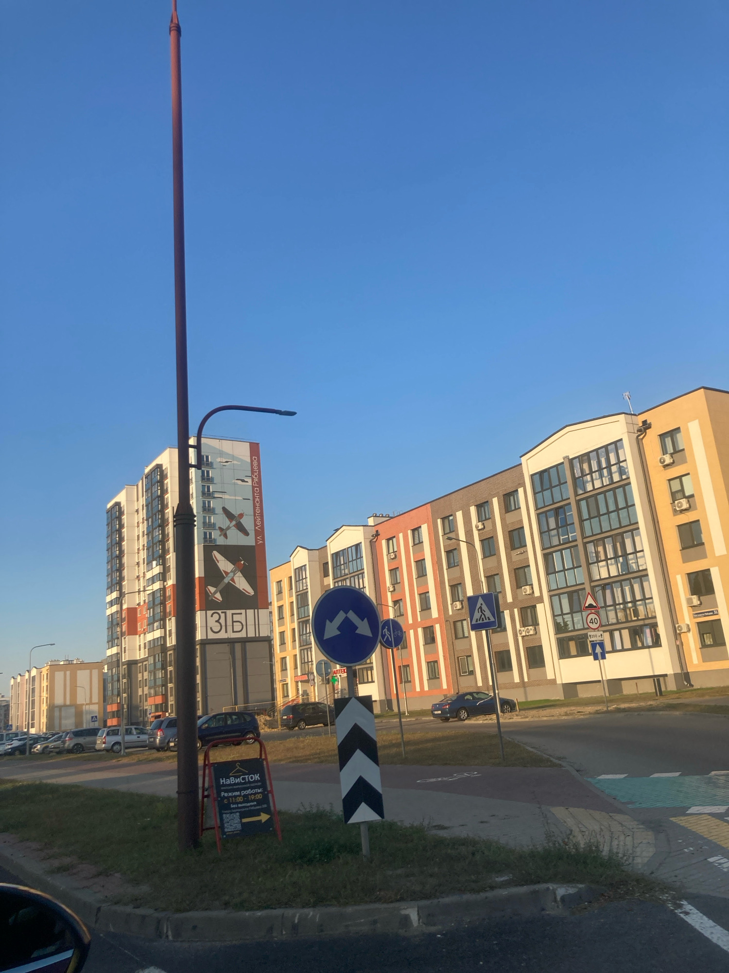The image depicts a street scene in Brest, Belarus. In the foreground, a street light pole stands to the left, with a white and red sign for "Navistok" on its base. The sign includes a QR code, operating hours (11:00-19:00), and a yellow arrow pointing right. To the right of the pole, traffic signs are visible, including a blue circular sign with arrows indicating a turn and a pedestrian crossing sign. Beyond the pole, a modern multi-story apartment building with colorful sections of orange, white, and gray is prominent. The building features many windows and air conditioning units. To the right of this building, another apartment building with yellow and white accents is visible. Cars are parked along the street in the mid-ground. The sky is clear and blue.