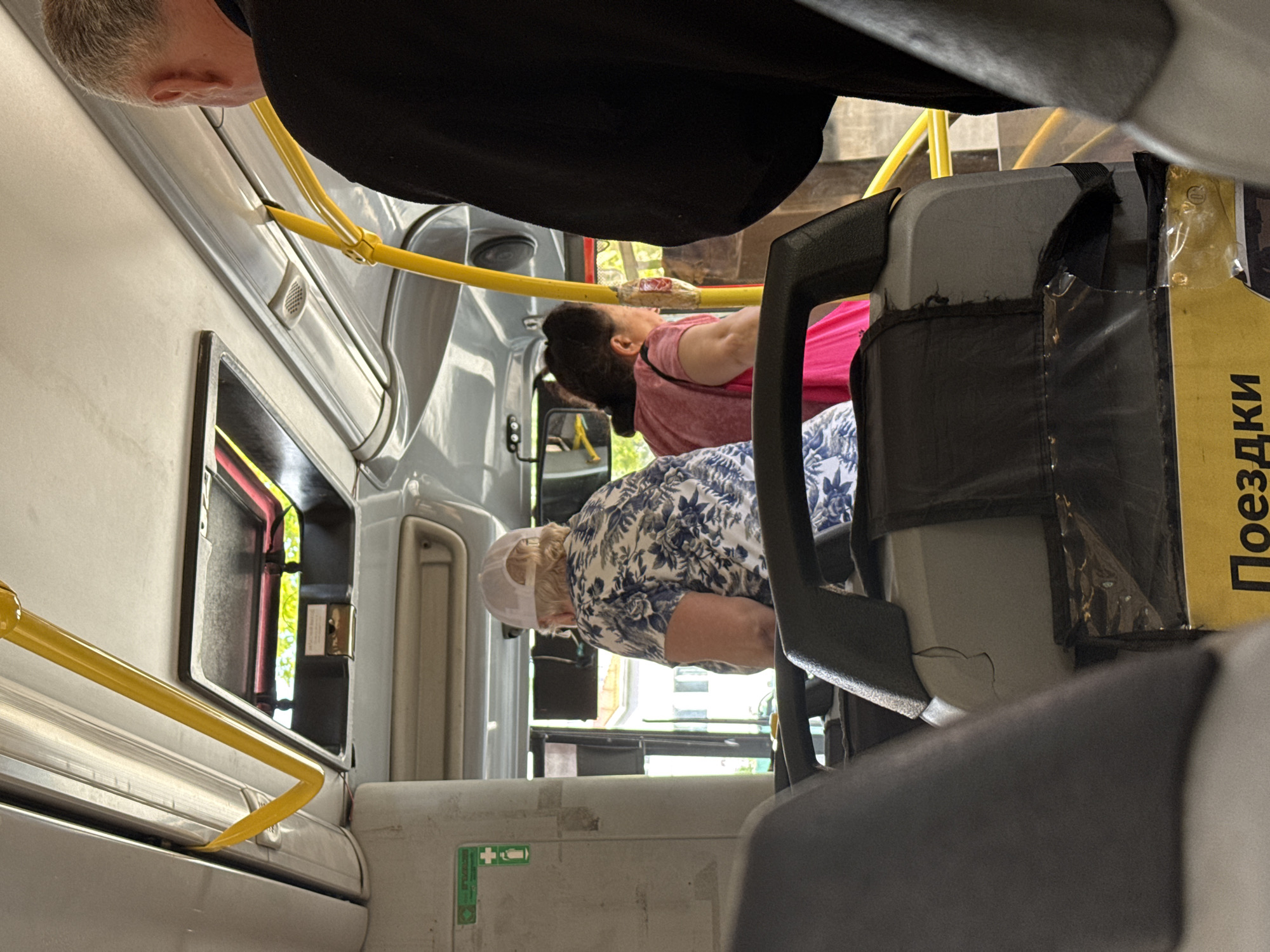 The image shows the interior of a bus, oriented sideways. In the foreground, a person with short, gray hair is leaning forward, their ear and the back of their head visible. They are wearing a black garment. Further into the bus, another person in a pink shirt is leaning forward, their back to the camera. They have dark hair pulled back. A third person, wearing a white cap and a floral-patterned shirt, is also leaning forward, their back to the camera. The interior of the bus is visible, with yellow handrails running along the top. Gray seats and parts of the bus interior are prominent. A yellow sign with black text is visible on the right side of the image. The lighting suggests it is daytime, with sunlight filtering in through the windows, illuminating parts of the bus and the scene outside. The overall atmosphere appears to be mundane, typical of daily public transportation.
The yellow sign on the right has Russian text that reads "Поездки" which translates to "Trips" or "Journeys". There is also a small green and white emergency exit sign visible on the lower left portion of the image.
