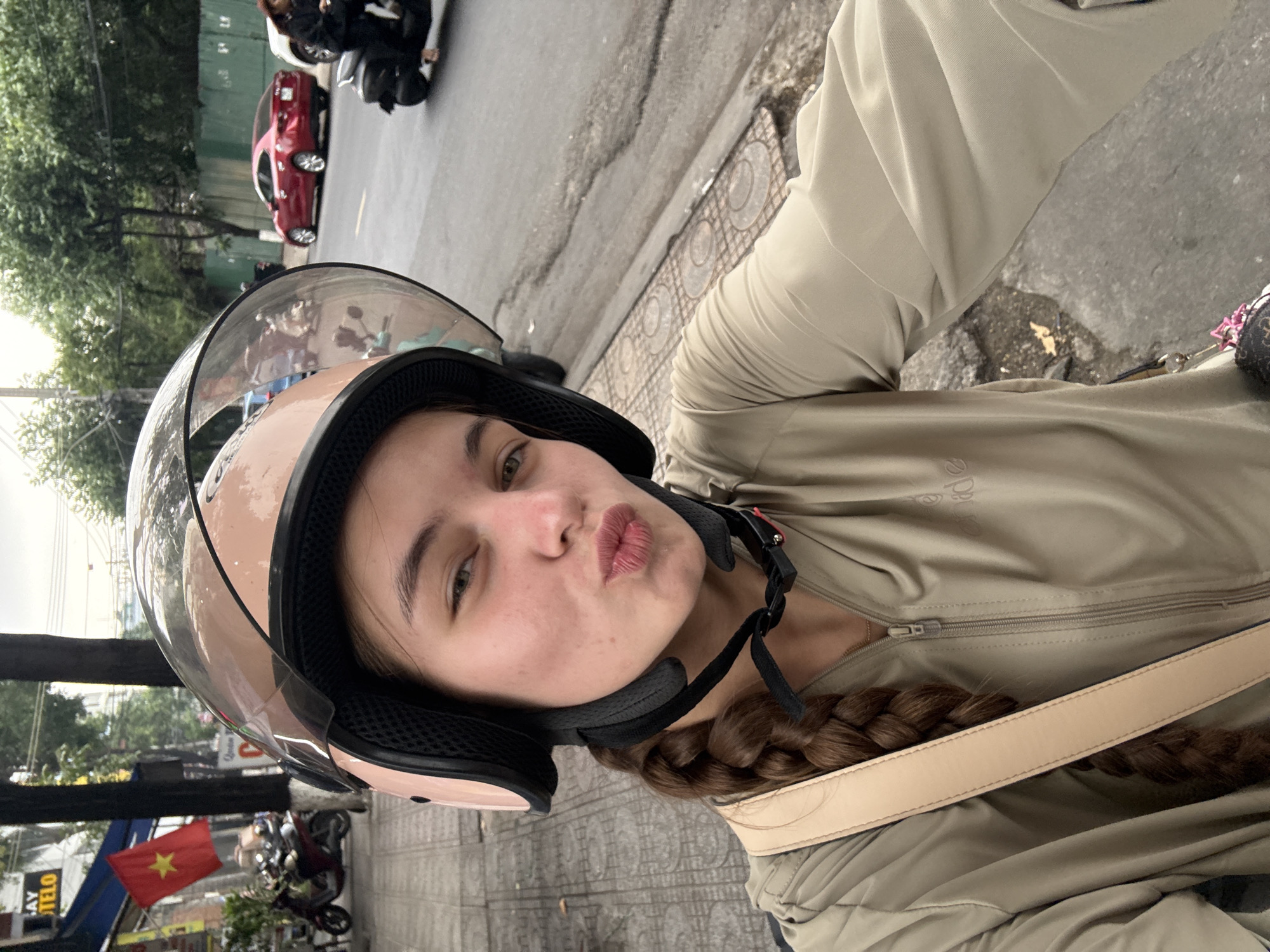 A young woman with a long braid, wearing a light tan jacket and a pink helmet, pouts at the camera. She is seated on a motorcycle, with the handlebars and dashboard partially visible. The background shows a city street with parked motorcycles, a red car, trees, and buildings. A Vietnamese flag is visible on a building to the left. The pavement is wet, suggesting recent rain.