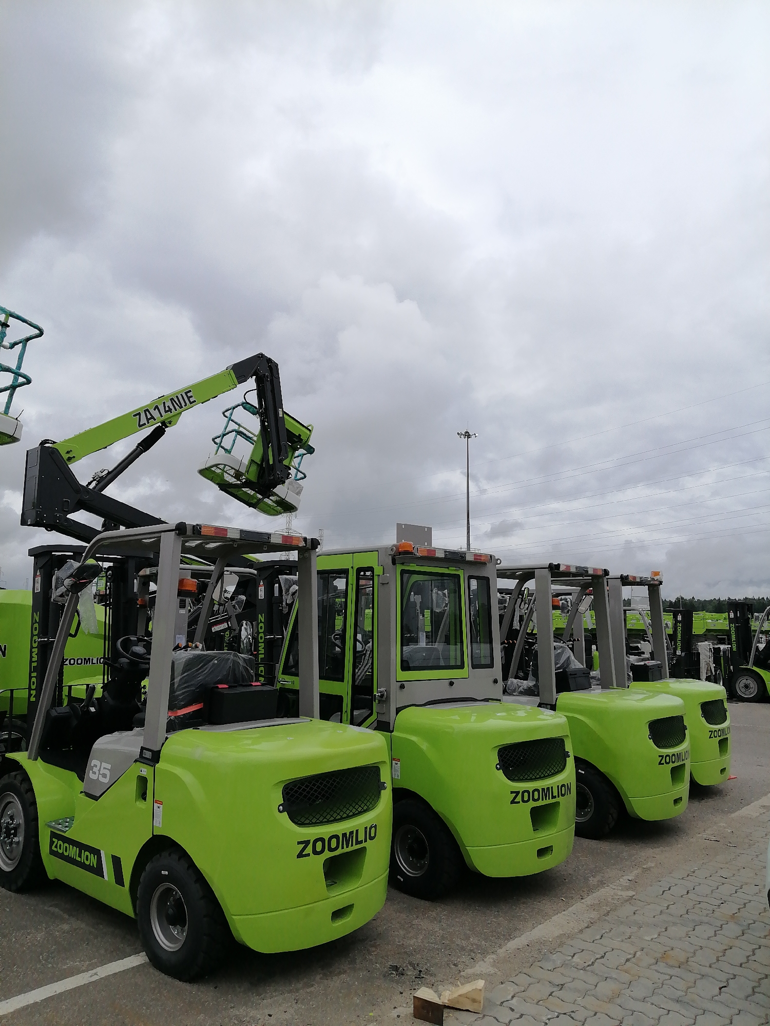 A fleet of bright green Zoomlion forklifts and an aerial work platform are parked on a paved lot under a cloudy sky. The aerial work platform, with a model number "ZA14NJE" visible on its boom, is positioned in the foreground, extending upwards. Several forklifts are lined up behind it, most with their "ZOOMILION" branding clearly visible. One forklift in the foreground has the number "35" on its side. In the background, more forklifts are visible, stretching into the distance. A light pole with several lights stands tall in the middle ground. The scene appears to be an industrial area or a storage yard. The location is specified as Спас-Каменка, Russia.