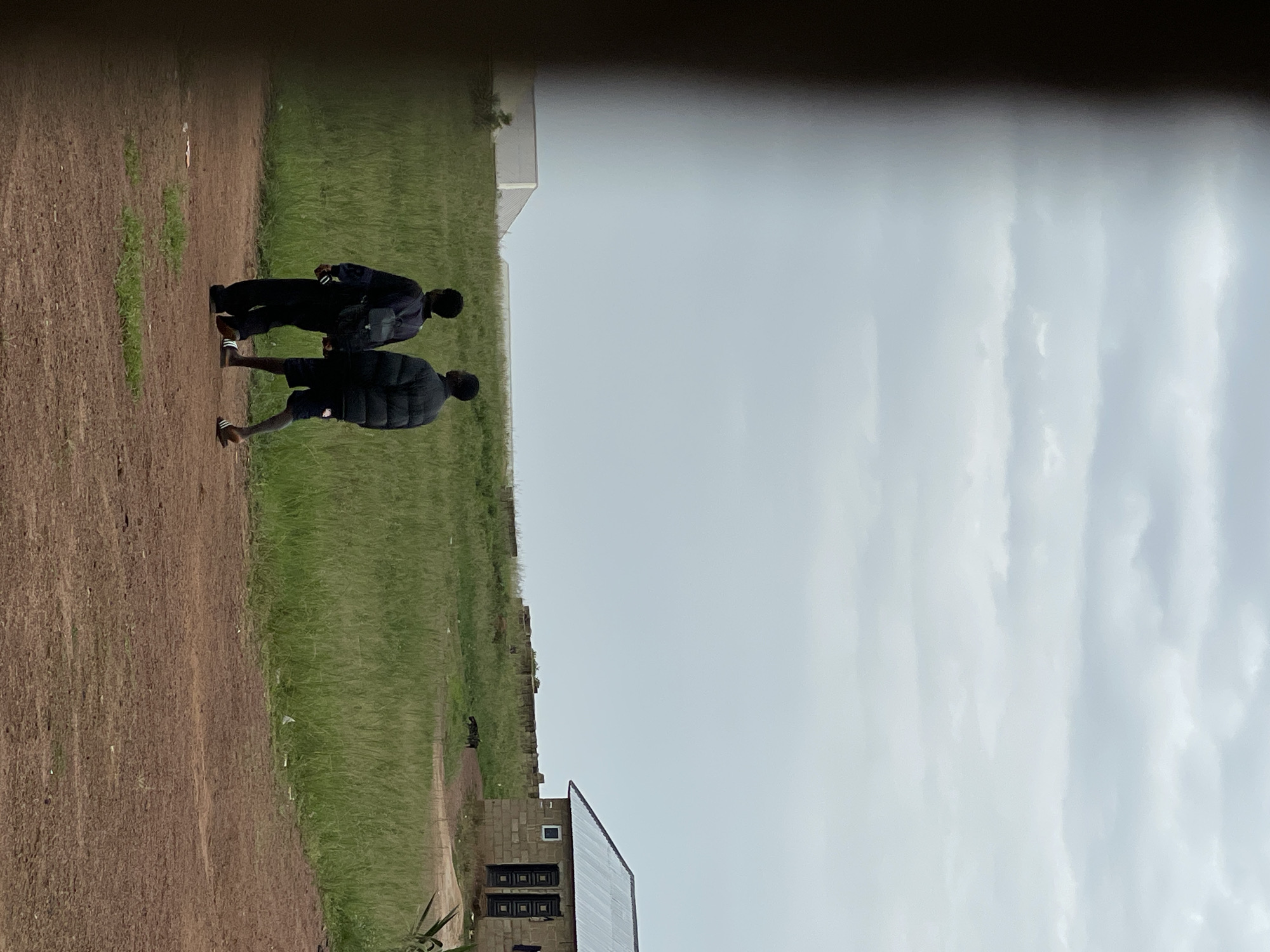Two young males, appearing to be teenagers, walk away from the camera on a reddish-brown dirt path in Durumi, Nigeria. The male on the left wears a dark blue jacket over a dark shirt and dark pants, with a black backpack on his shoulders. The male on the right wears a dark puffer jacket and dark shorts, with dark sandals. They are walking in a grassy field under an overcast sky. In the background to the right, a small, single-story building constructed of concrete blocks with a corrugated metal roof is visible. The building has two doors with dark frames. Further in the background, a stone wall runs across the landscape, and two dogs can be seen near the building and the wall. To the left of the frame, a blurred, dark object partially obstructs the view of the scene. Industrial-looking white structures are visible on the horizon in the far left of the image.