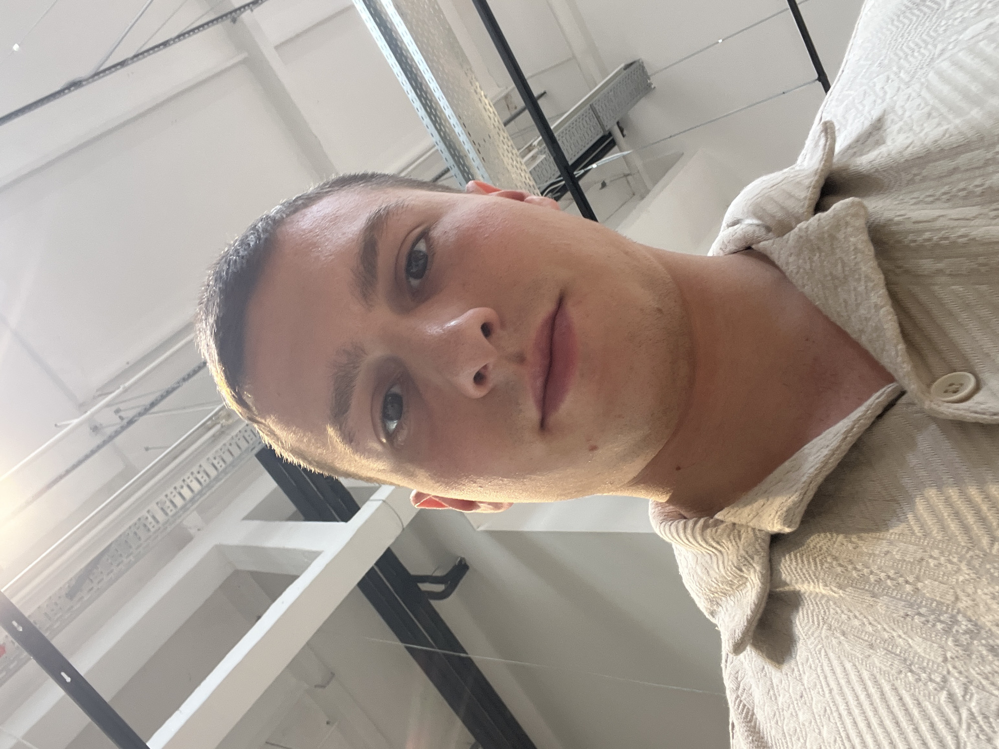 A low-angle, close-up photograph captures a man's face and upper torso against a backdrop of an industrial-looking ceiling.
The man has short, light brown hair, light skin, and appears to be in his 20s or 30s. He has a neutral expression and is looking directly at the camera. He is wearing a beige, textured shirt with a visible button.
The background reveals a commercial or industrial space. It features exposed pipes, beams, and metal grating. The lighting appears to be bright and somewhat diffuse.
It is impossible to determine if the location is in Varna, Bulgaria, from the image alone.