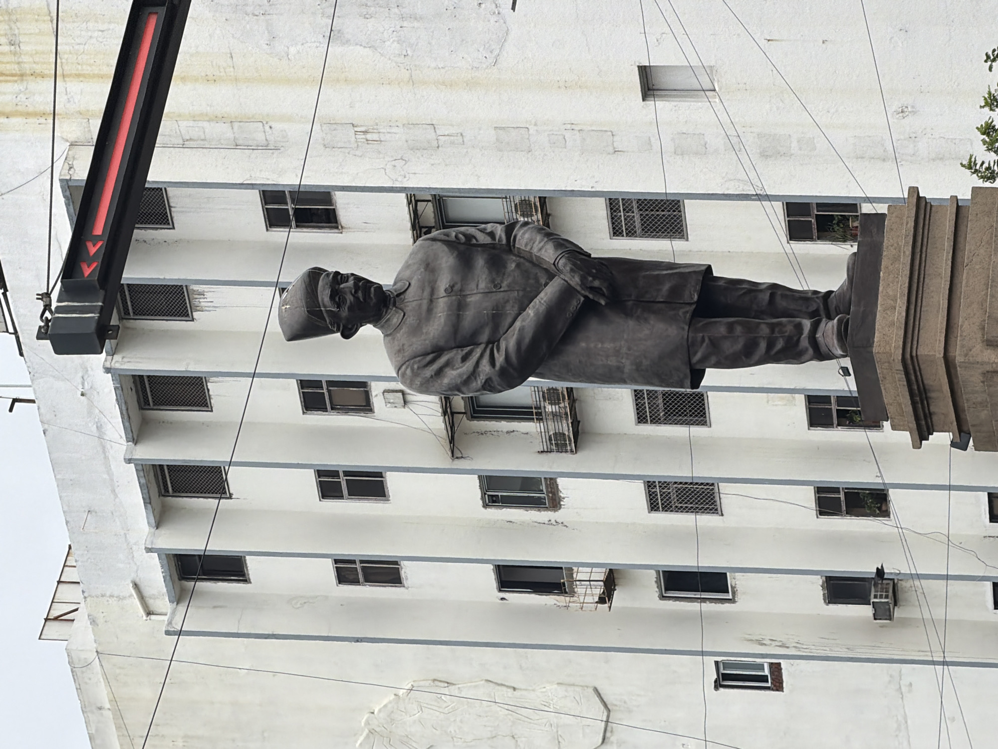 The image shows a bronze statue of a man in a traditional cap and coat, standing on a stone pedestal in front of a white, multi-story building. The building has many windows, some with metal grates. There are also electrical wires crisscrossing the scene. Above the statue, a black metal beam with red arrow indicators is visible. The statue appears to be in a public space, possibly a city square or park. The building's architecture suggests it is an older structure. The overall impression is one of a monument or memorial in an urban environment. This statue is located at Dhobi Talao, Mumbai, India.