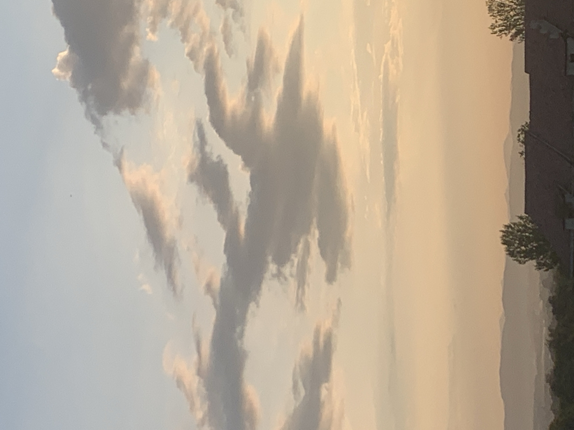 This is a photograph of a cloudy sky over a landscape. The sky is a gradient of blue and pale orange, with several clouds scattered throughout. Some of the clouds are dark gray, while others are tinged with pink and yellow from the setting sun. In the distance, there are hazy mountains and hills. In the foreground, the rooftops of houses and trees are visible. The image does not contain any identifiable people or specific landmarks that would indicate the location in Kolomyia, Ukraine. The scene appears to be taken during dusk or dawn.