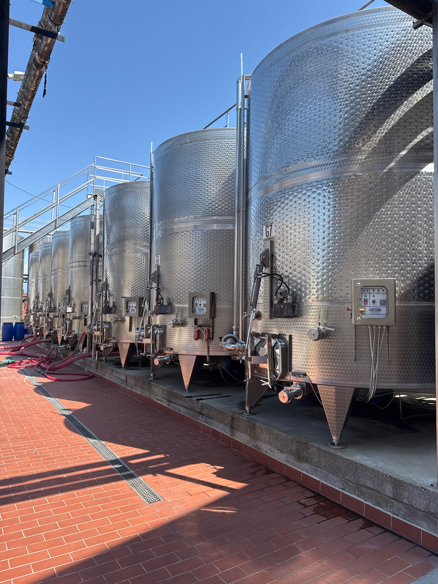 The image depicts a row of large, stainless steel fermentation tanks, likely at a winery, situated outdoors under a clear blue sky. The tanks are cylindrical with a textured surface and are supported by conical legs. Various pipes and control panels are attached to the sides of the tanks. In the foreground, a red brick paved area with a drainage grate runs alongside the tanks. Red hoses are visible, connected to the tanks and lying on the ground. Two blue barrels are also present near the leftmost tanks. The scene appears to be in an industrial or agricultural setting, possibly a wine production facility. Based on the location provided, this scene is in Kurdghelauri, Georgia.
