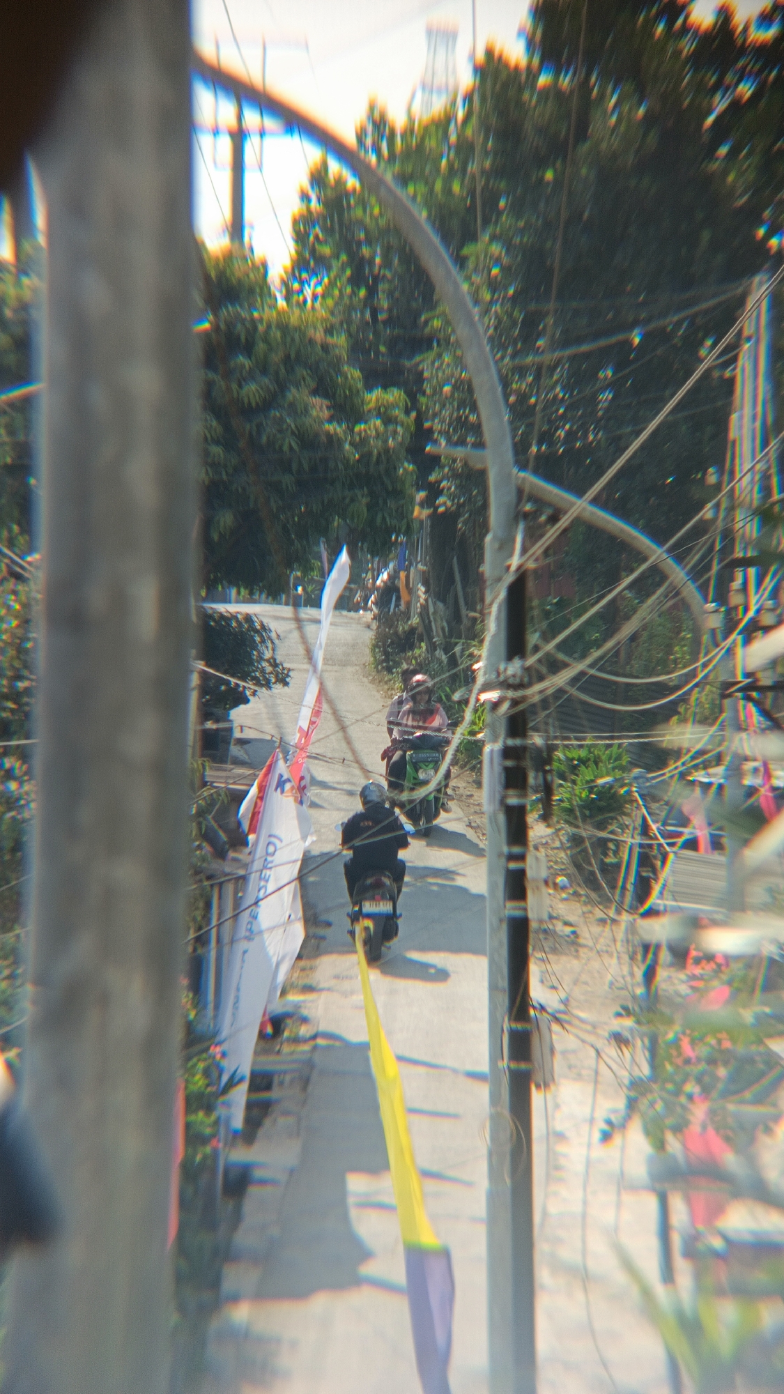 A long, narrow, paved road winds uphill through a green, leafy environment. The road is lined with trees on both sides, and power lines crisscross the scene overhead. Two motorcycles are visible on the road, both traveling uphill. The lead motorcycle is green, ridden by a person wearing a red shirt and a dark helmet. Behind them, a dark motorcycle is ridden by a person wearing a black jacket and a helmet. Several flags are also present along the side of the road. One flag, white with red lettering, is prominent near the foreground, partially obscuring the view of the motorcycles. Another flag, yellow and purple, is visible further down the road. The overall scene appears to be in a semi-urban or rural area of Margajaya, Indonesia, likely on a sunny day given the strong shadows.