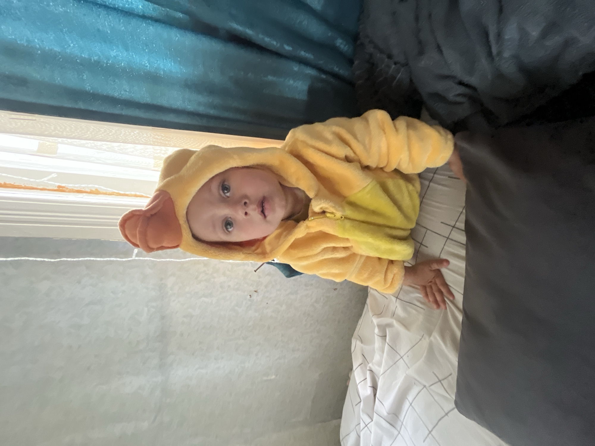 A young child, appearing to be a toddler, is lying on a bed, looking up at the camera. The child is wearing a yellow hooded costume, possibly a duck or chick, with orange ear-like details on the hood. The background consists of blue curtains and a window, with white bedding on the bed. The scene is indoors. The lighting suggests it could be daytime. The child's expression is neutral, with wide eyes looking towards the viewer. The image is oriented vertically, with the child's head towards the top left.